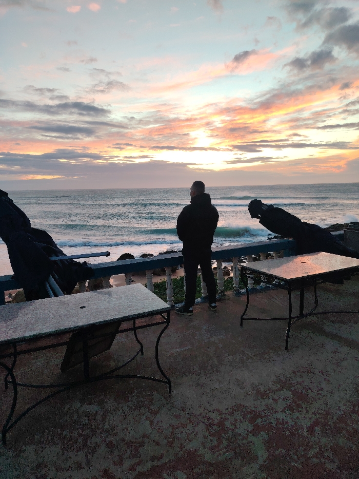 The image displays an outdoor terrace scene at twilight, overlooking the sea in Tanger, Morocco. A single male subject stands with his back to the camera, observing the ocean. He is dressed in a dark, possibly hooded, jacket, dark trousers, and athletic shoes. He stands behind a blue railing with white balusters separating the terrace from the sea. The ocean exhibits moderate waves breaking against a visible rocky shoreline. The sky above is characterized by twilight colors, featuring hues of pink, orange, purple, and blue, with scattered clouds. The foreground terrace is made of concrete and shows patches of discoloration. Two square tables with light-colored tops and dark, ornate metal bases are positioned on the terrace. A dark, covered object, possibly a folded chair, is present near the table on the left. A second similar dark object is partially visible to the right of the standing subject, positioned behind the rightmost table.