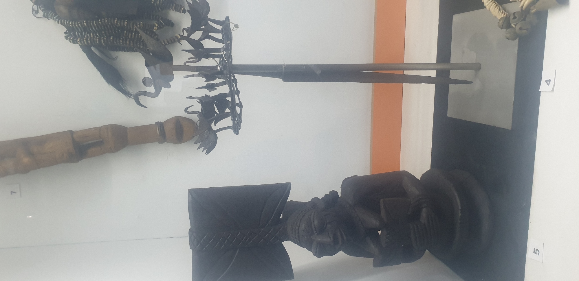 The image shows an interior display, possibly in a museum, presenting cultural artifacts. In the foreground is a dark wooden carving of a figure with a stylized axe. Above it is a collection of objects, including a spear-like item with a metal shaft and a decorative, possibly ceremonial, headdress or adornment. A wooden handle is visible beside the headdress, possibly related to the artifacts. The number tags "4", "5", and "7" are visible next to the artifacts, which likely identify them. The location is Vom, Nigeria. The items are positioned against a white and orange wall.