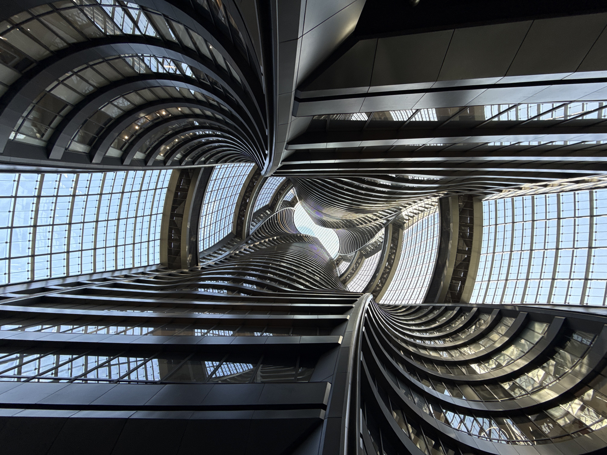 The image is a low-angle view of the interior of a modern building. The architecture is characterized by sweeping curves and lines, creating a vortex-like effect towards a central skylight. The building's structure is predominantly constructed of metal and glass, with horizontal lines suggesting multiple levels or floors. The glass allows natural light to permeate the interior. The scene is located in the Fengtai District of China.