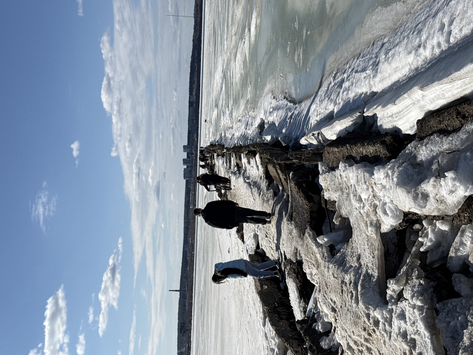 The image depicts a partially frozen body of water with a snowy shoreline, possibly a lake or river. In the foreground, three people are walking along the shoreline. One person is wearing a dark jacket and pants, while another is wearing a blue jacket and jeans. A third person appears to be riding a bicycle further down the shoreline. The shoreline is composed of dark rocks and snow. In the background, there is a cityscape with a tall tower visible. Based on this tower and the general landscape, this is a plausible depiction of Izhevsk, Russia. The weather appears to be sunny with some clouds in the sky. The water is partially covered in ice, indicating it is likely winter or early spring.