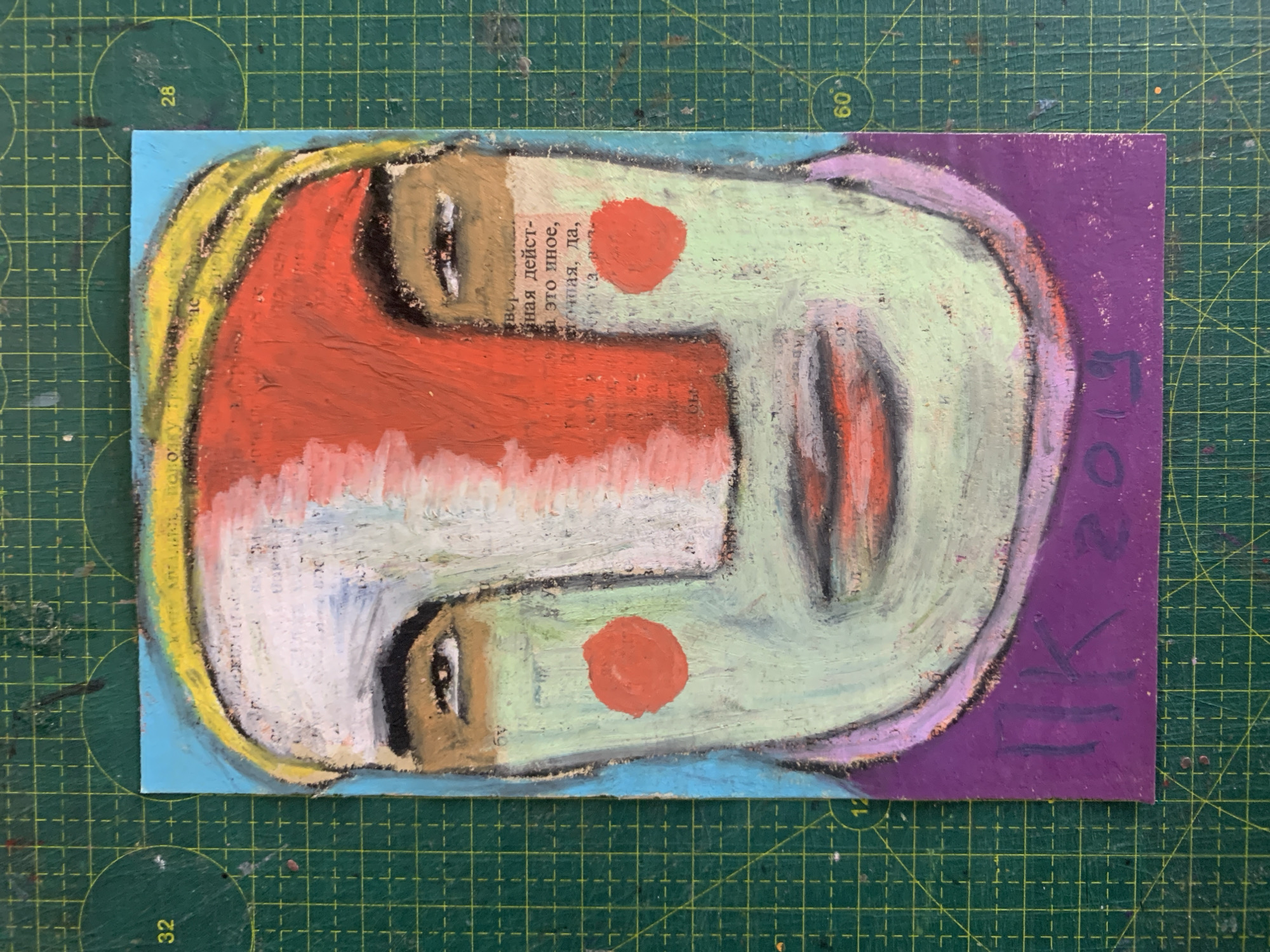 This is a photograph of a mixed-media artwork, likely a portrait, displayed on a green cutting mat. The artwork itself is a vertical rectangle featuring a stylized depiction of a face. The face is rendered in a primitive, almost cubist style with a light blue background and a yellow band across the top of the head. The skin tones are predominantly pale green with a prominent red area on the forehead and cheeks. The eyes are almond-shaped, and the mouth is full and red. There are two circular orange marks on the cheeks, resembling blush or decorative elements. Text from a newspaper or book is visible collaged onto the face, particularly on the forehead and cheeks. The artwork has a purple border at the bottom, upon which is written in purple capital letters, "HK 2019". The cutting mat has a grid pattern with circular markings indicating measurements, including "32", "28", "12", and "60". The overall impression is that of an artist's studio or workspace, with the cutting mat serving as a backdrop. There is no discernible connection to the city of Sergeevo, Russia, in the image itself.