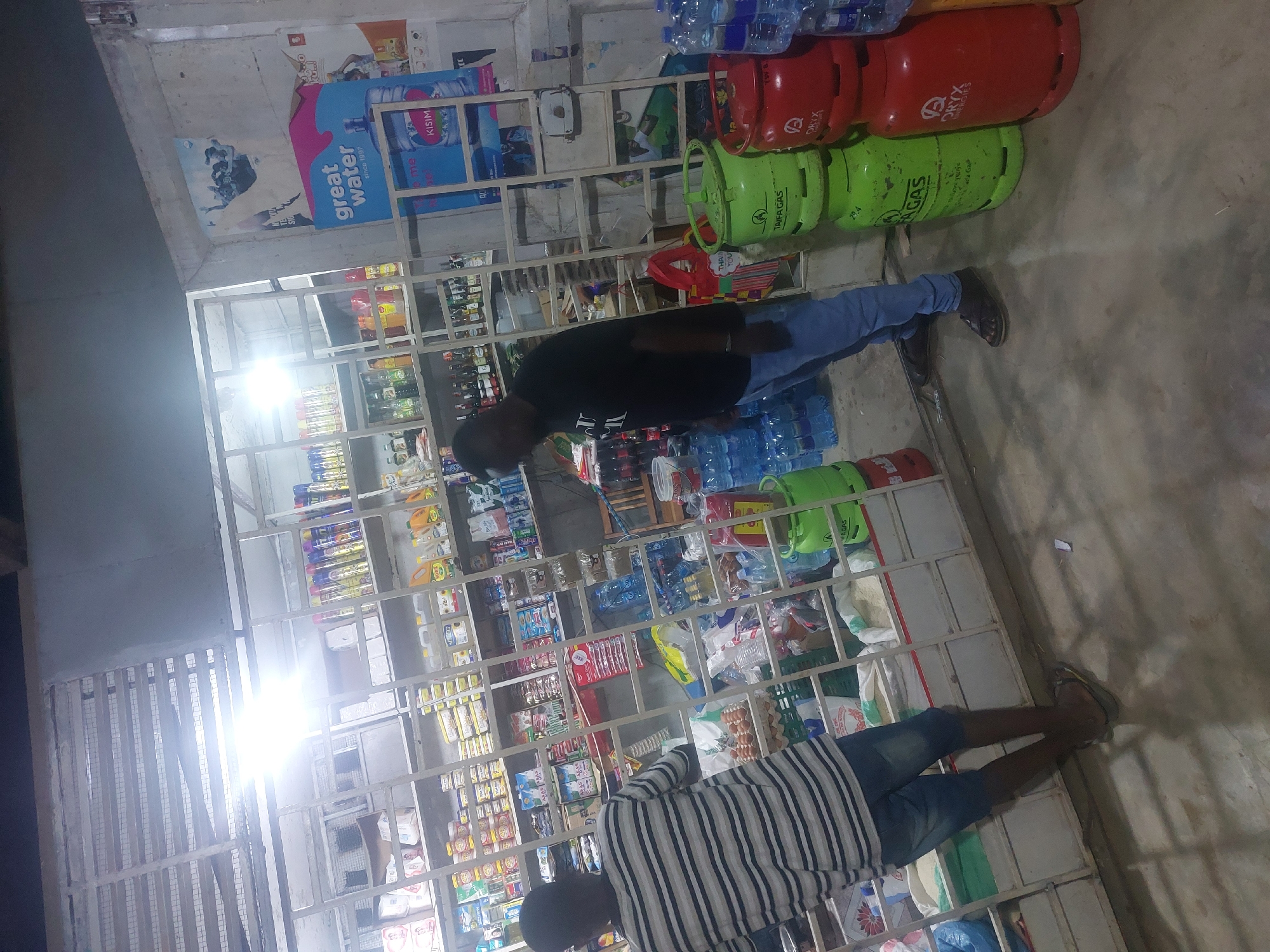 The image shows the interior of a small store, likely in Dar es Salaam, Tanzania, given the brands of gas cylinders and water bottles visible. Two men are visible in the store. One is wearing a black t-shirt and jeans and appears to be examining items on the shelves. The other man is wearing a striped shirt and jeans, also looking at items on the shelves. The store is well-stocked with a variety of goods, including drinks, food items, and gas cylinders. The shelves are made of metal and are organized in a grid pattern. The lighting in the store is bright. Gas cylinders, some red with the "ORYX" logo and some green with the "TAIFA GAS" logo, are stacked on the top shelves. Bottled water with the "Great Water" brand is also prominently displayed.