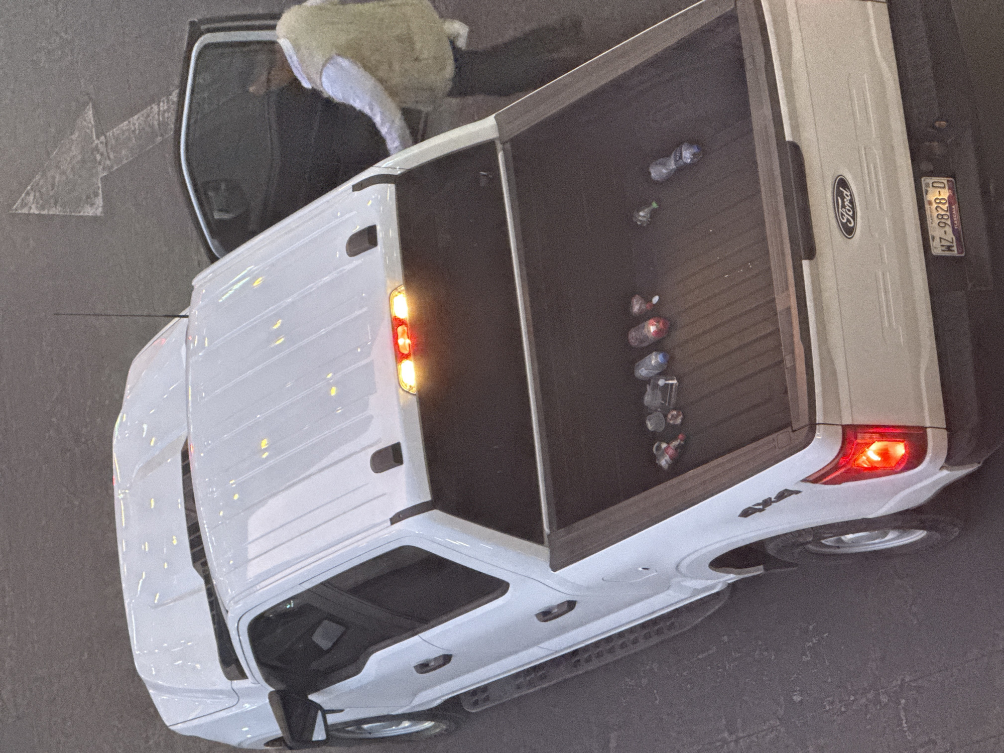 A top-down view shows a white Ford pickup truck parked on asphalt. The truck's tailgate is down, revealing several empty water bottles and a small bottle inside the bed. A person is partially visible near the open driver's side door. An arrow marking a traffic lane is painted on the asphalt near the front of the truck. The license plate reads "WZ-9828-D" and indicates Tlaxcala. "4x4" is printed on the rear of the truck. The scene is located in San Bernardino Tlaxcalancingo, Mexico.