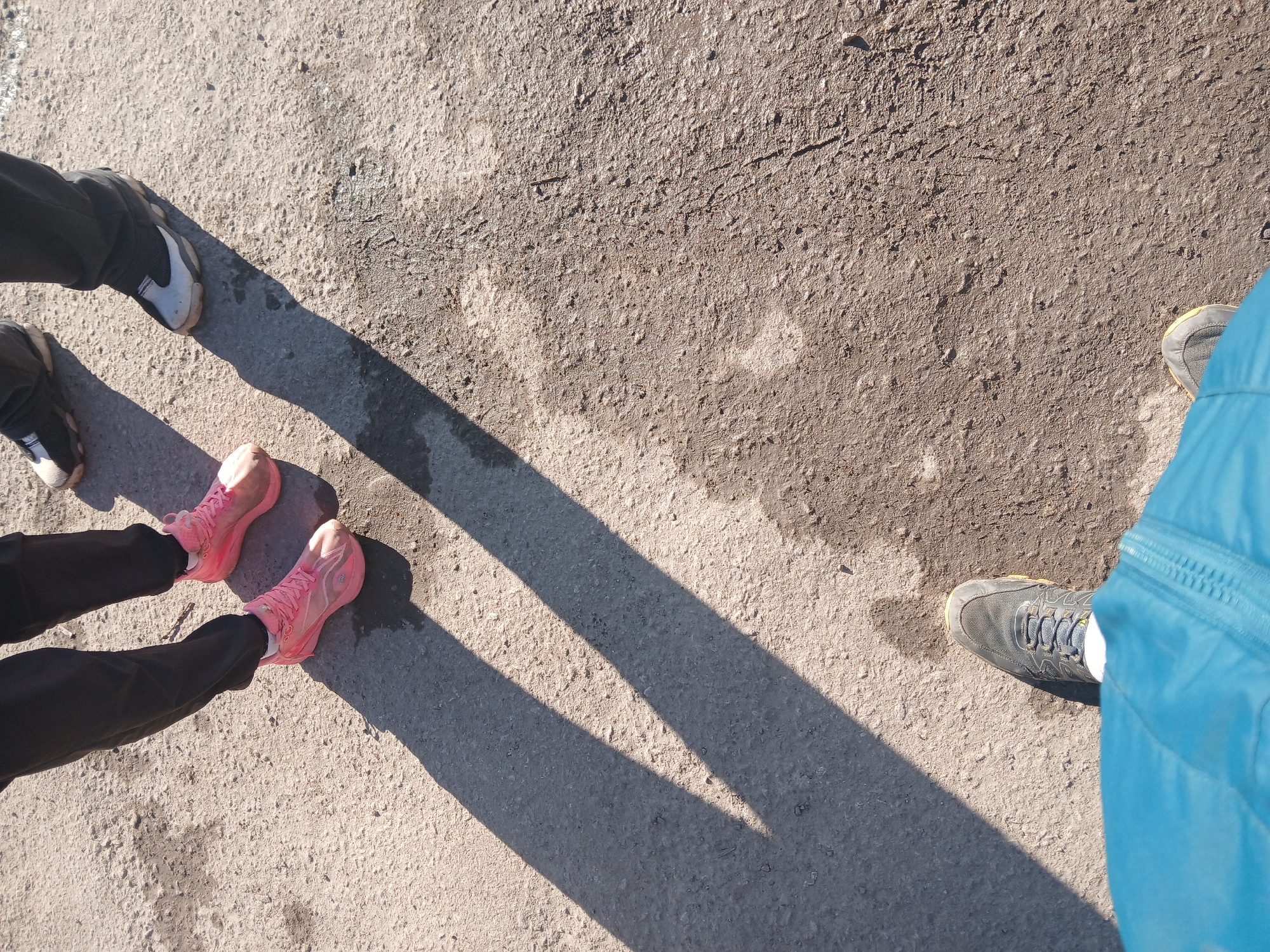 The image shows a concrete surface with three pairs of shoes and their shadows. In the lower left, a person is wearing black pants and pink athletic shoes. Near the upper left corner, a person wears black pants and black and white sneakers. In the lower right corner, a person is partially visible wearing blue pants and gray hiking shoes. The concrete surface is uneven and textured, with variations in color.