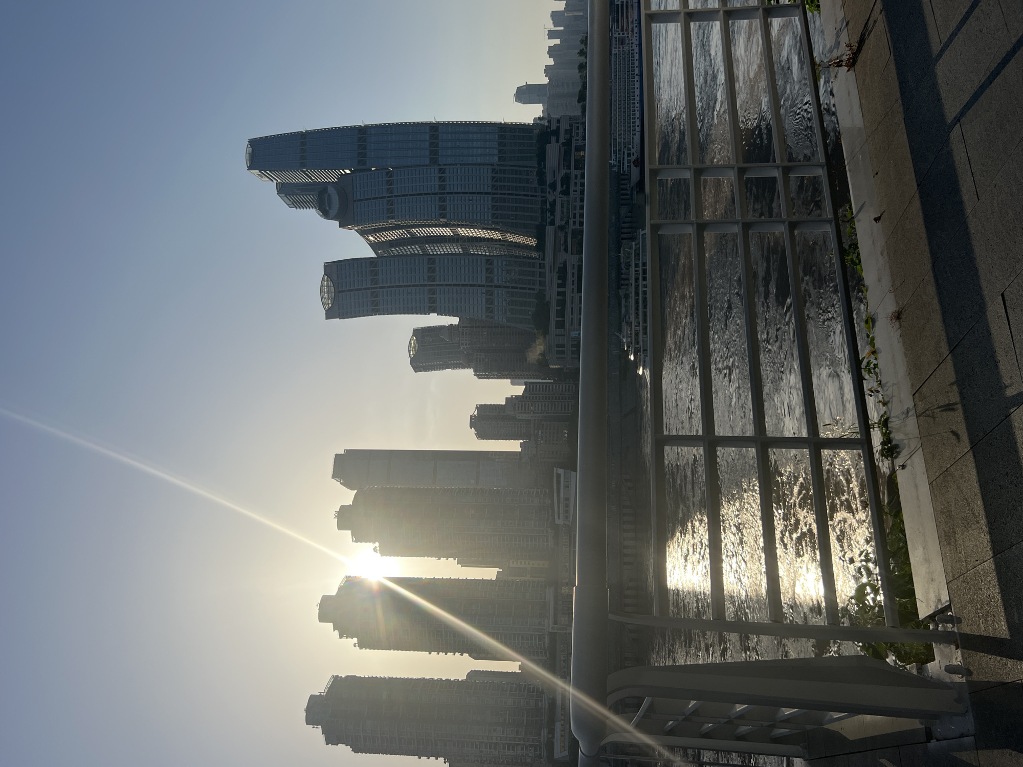 The image captures a panoramic view of the Nan'an District in Chongqing, China, during what appears to be sunrise or sunset. The sun is low in the sky, creating a bright glare that obscures some of the detail of the modern skyscrapers in the background. These buildings are a prominent feature of the skyline, with some appearing to be under construction. In the foreground, a railing separates a paved walkway from a wide river. The river's surface reflects the sunlight, creating a shimmering effect. Two large passenger boats are visible on the water. The walkway is made of stone tiles, and shadows from the railing stretch across it. There are no people visible in the image. The sky is clear and blue, with a slight haze in the distance.