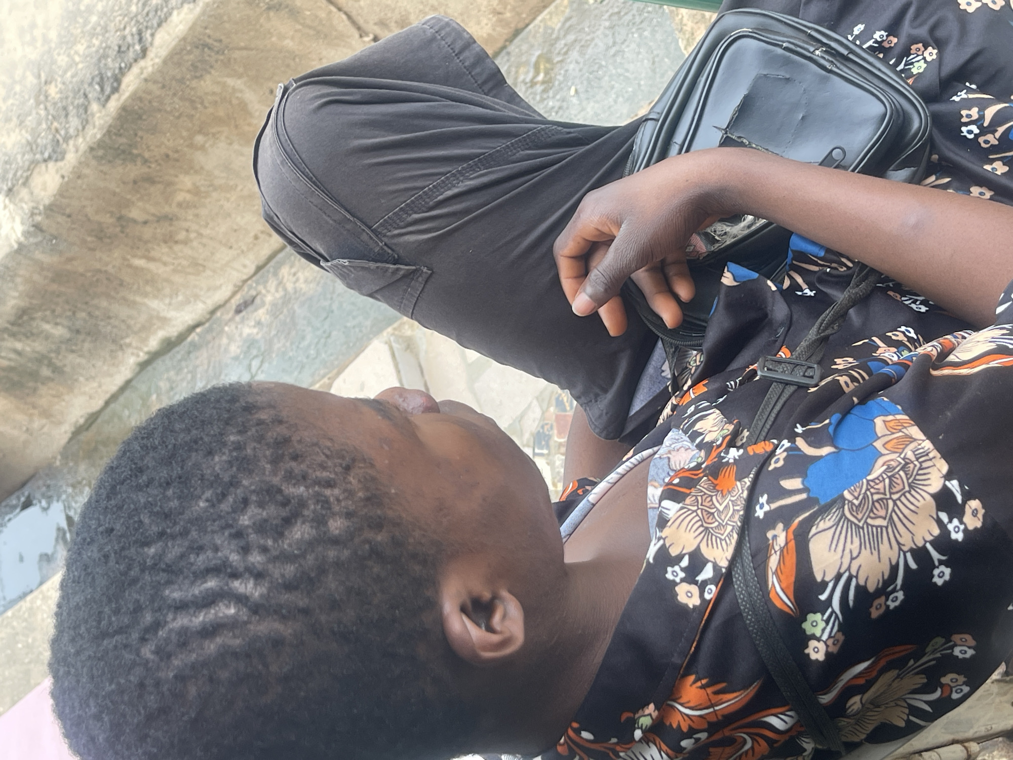 A person, likely a young woman, is sitting outdoors on a concrete bench. She has dark skin and short, dark hair styled in waves. She is wearing a black dress with a floral print featuring orange, blue, and beige flowers. A black crossbody bag with a zippered compartment rests on her lap. Her left hand is visible, resting on her leg. The background consists of weathered concrete structures and some water, suggesting an outdoor public space. The scene is captured in a medium shot, focusing on the individual and her immediate surroundings.