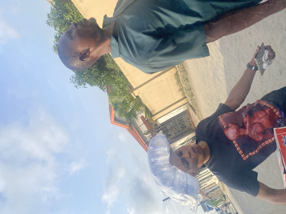 The image depicts an outdoor daytime street scene in Iranla, Nigeria. In the foreground, a dark-complexioned woman is captured from the chest up, positioned slightly left of center, wearing a black short-sleeved t-shirt with a graphic of a shirtless male figure. She carries a clear plastic bag containing multiple sachets of "PLOM" brand drinking water balanced on her head. In her left hand, she holds a red and white box, identifiable as "Indomie" noodles. Her expression is focused, and her gaze is directed slightly downwards and to her right. To the right and slightly behind the woman, a dark-complexioned man is seen in profile, looking upwards and to the right. He wears a dark teal short-sleeved collared shirt and exhibits visible perspiration on his face and arm. In his left hand, he holds a small, dark, folded object. The background reveals a light-colored, unpaved road, a tan-colored wall, green foliage including trees and bushes, and a building with an orange-red gabled roof and a dark metal gate. The sky is light blue with scattered white clouds. Power lines are partially visible on the far left. The two individuals appear to be in transit, but no direct interaction between them is evident.
