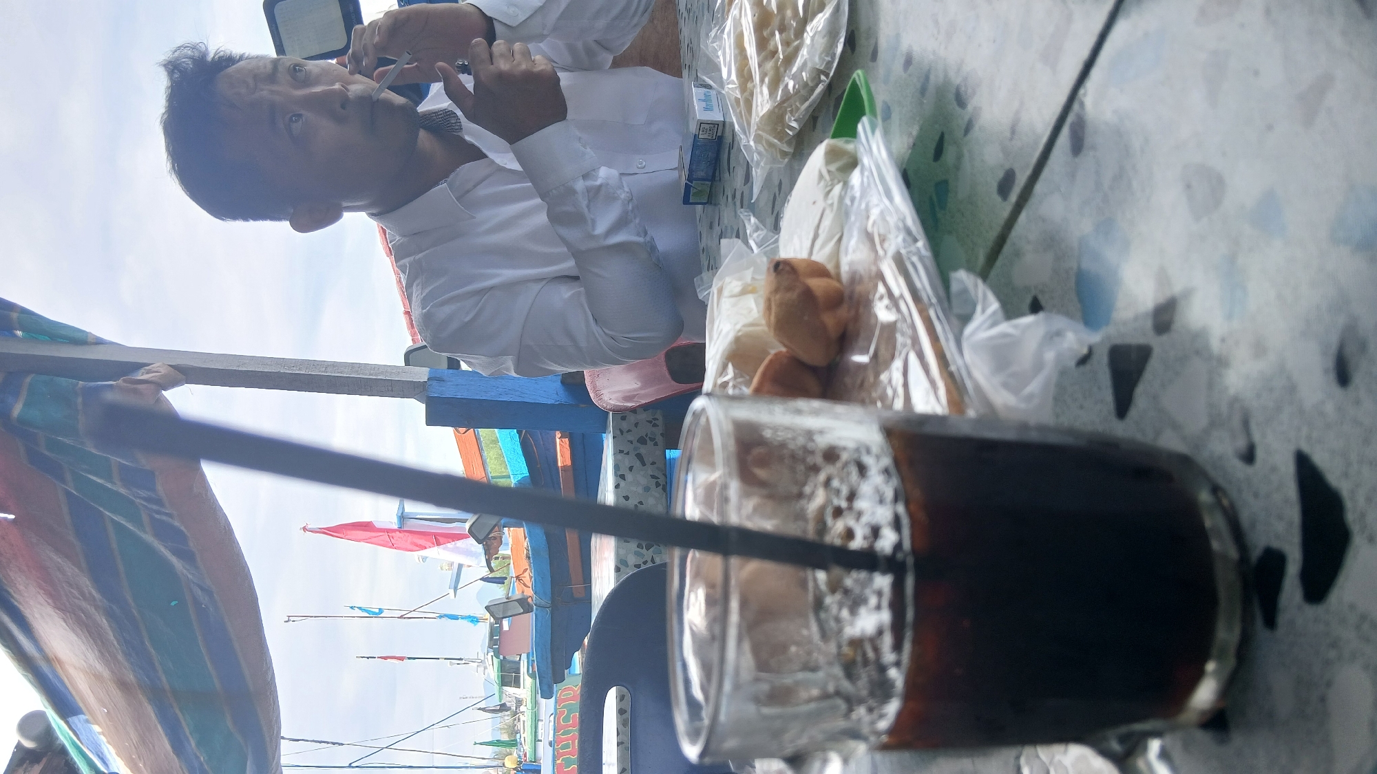 In Banda Aceh, Indonesia, a man in a white shirt is seated at a table outdoors, with boats docked in the background under a bright, cloudy sky. He is in the process of lighting a cigarette, holding a lighter in his right hand and the cigarette in his left. In front of him on the table, there is a glass of dark liquid with a black straw, a pack of Marlboro cigarettes, and some wrapped food items, possibly pastries. The table has a speckled surface with shades of gray, white, and blue. The scene appears to be near a harbor or waterfront, suggested by the presence of fishing boats and a canopy overhead.