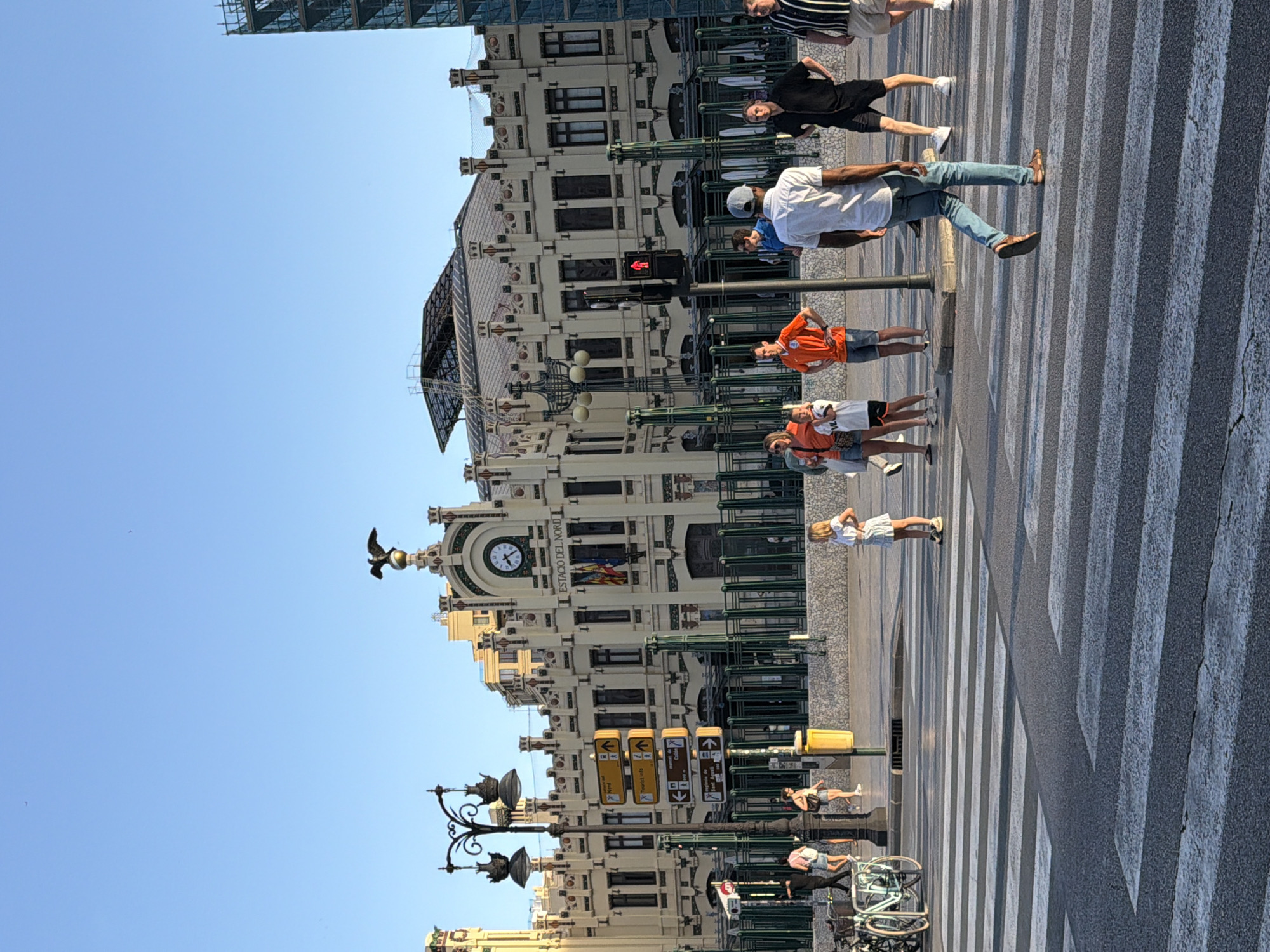 In front of the Estació del Nord, a historic train station in Valencia, Spain, people are crossing a street at a pedestrian crossing. The station, a prominent building with ornate architecture and a large clock, serves as the backdrop. Several individuals are captured in motion, some walking across the zebra stripes, while others are standing or conversing on the sidewalk. The sun is casting long shadows, indicating it is likely late afternoon or early morning. The scene is bustling with activity, typical of a public space in a city.