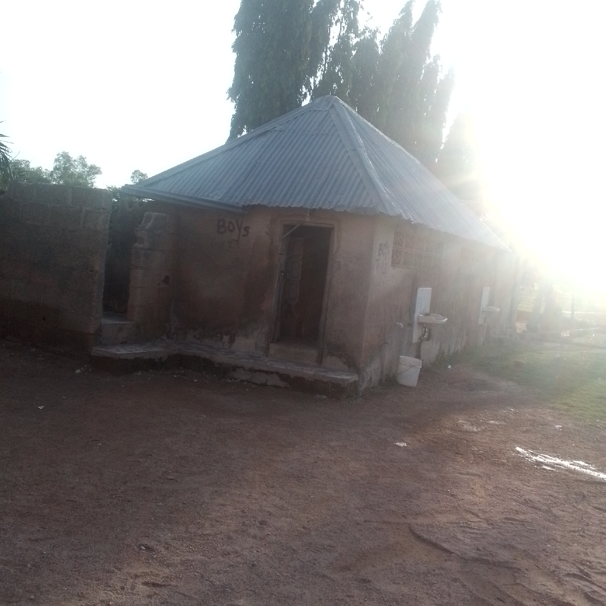 The image depicts the exterior of a building, likely a public restroom, in Jalingo, Nigeria. The building has a corrugated metal roof and walls constructed from mud or a similar earthy material. The entrance to the building is visible, with steps leading up to it. On the wall to the left of the entrance, the word "BOYS" is spray-painted, followed by "TOILE". Another similar inscription is visible on the wall to the right of the entrance. Two white sinks are mounted on the exterior wall of the building, one on each side of the entrance, with a white bucket placed beneath the one on the right. A few scattered pieces of trash are visible on the ground. Tall trees are visible in the background, and the sky is bright with a strong glare from the sun, suggesting it is late afternoon or early morning. The overall setting appears to be somewhat rural or undeveloped.