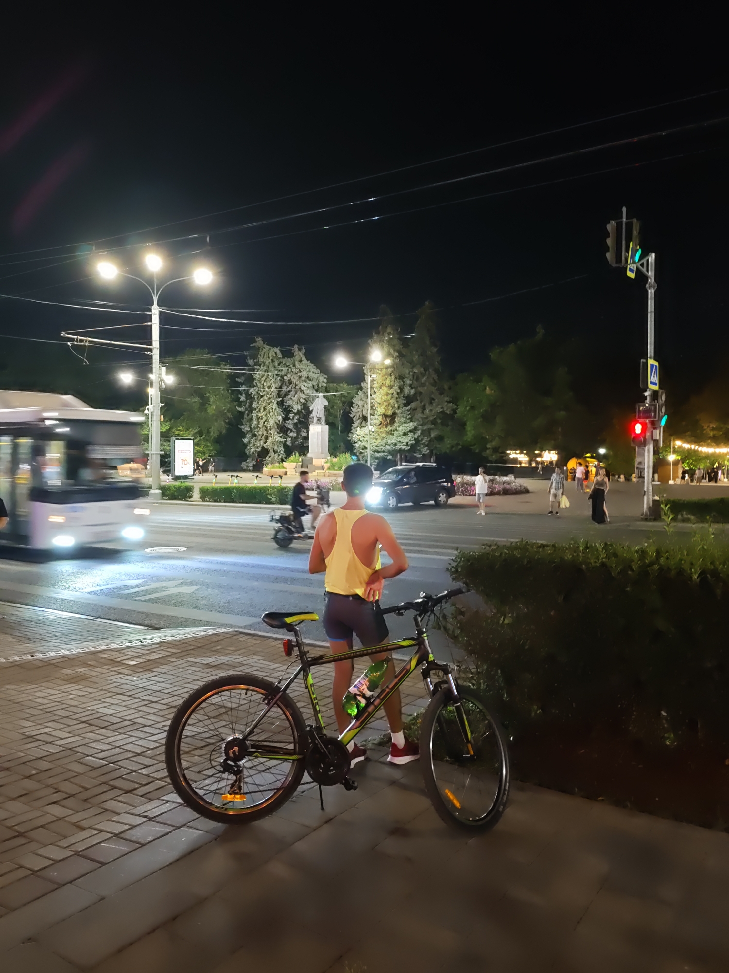 The image depicts a nighttime urban scene. In the foreground, a bicycle is parked on a brick-paved sidewalk. Behind the bicycle, a person wearing a yellow tank top and dark shorts stands facing away from the camera, looking towards a street. The street has a crosswalk and appears to be moderately busy, with a bus in motion on the left side of the frame. Further in the background, there are trees, streetlights, a traffic light displaying red, and several other pedestrians. The environment suggests a public area within a city, possibly a park or a promenade, illuminated by artificial lights.