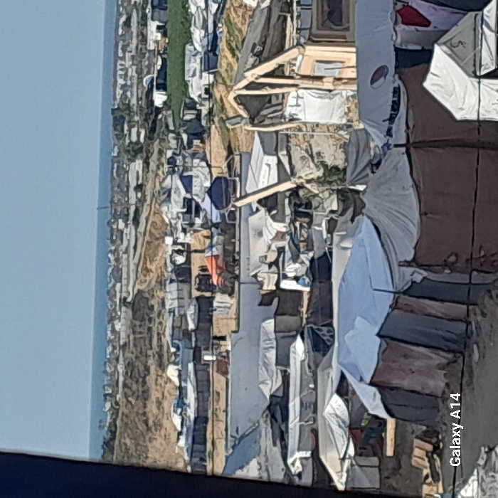 The image displays a sprawling, densely packed displacement camp in Khan Yunis, Palestinian Territory, under clear daylight conditions. Numerous temporary shelters and tents, constructed from various materials including white, light blue, grey, and brown tarpaulins and fabrics, dominate the foreground and midground. Some structures incorporate visible wooden frames. The camp is situated on sandy, undulating terrain, with areas of cleared ground and low concrete walls visible. In the midground, a prominent orange tarp-covered object is discernible among the shelters. Beyond the immediate settlement, the landscape transitions into sandy hills, which meet a coastal strip featuring scattered low-rise buildings. The Mediterranean Sea stretches across the distant horizon, beneath a clear blue sky. No specific individuals, actions, or interactions are clearly identifiable within the frame due to the distance and resolution.