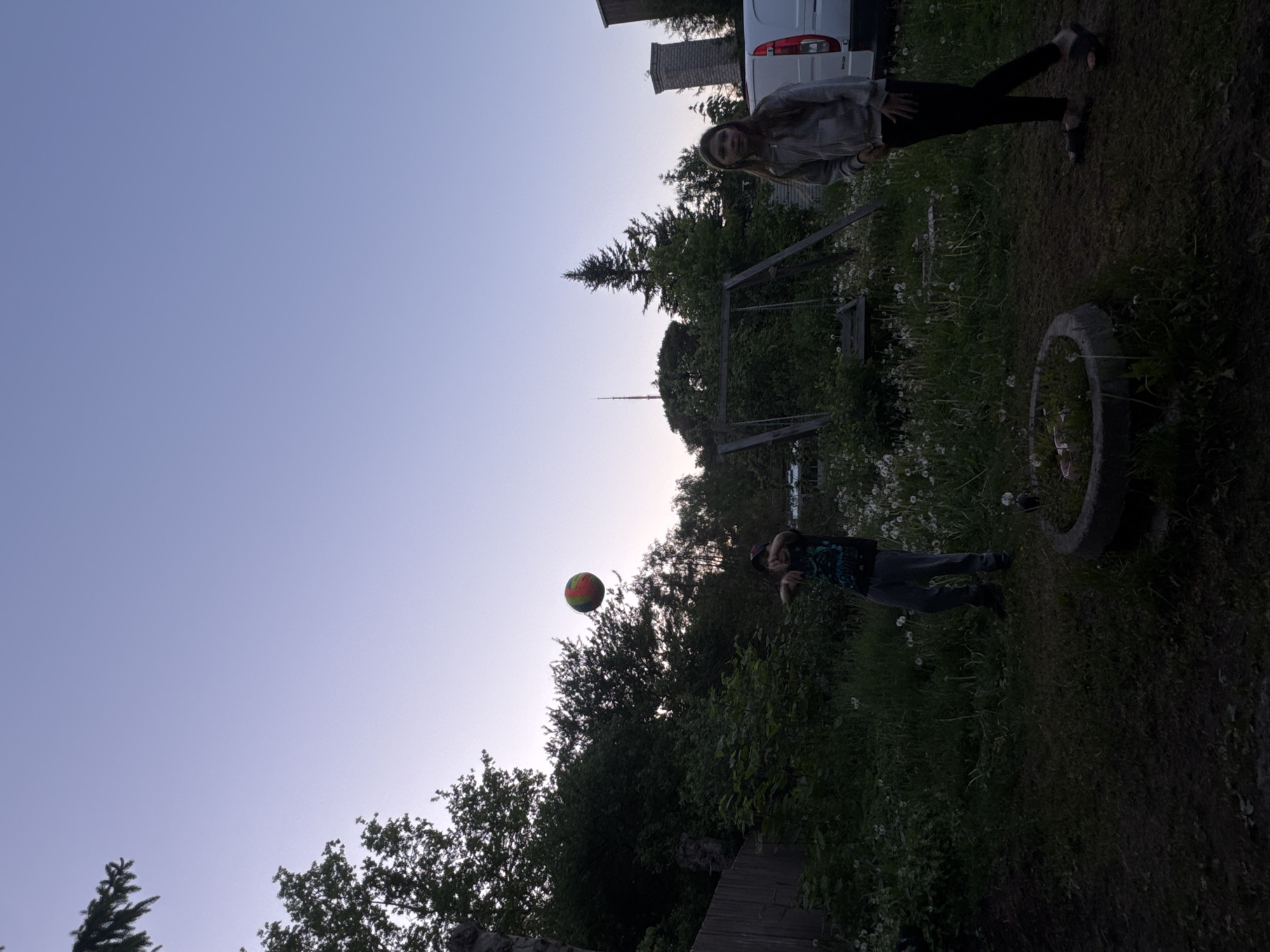 The image depicts a residential backyard scene at dusk in Tallinn, Estonia, as identified by the visible Tallinn TV Tower in the distant background. Two individuals are present. On the left, a person wearing a dark graphic t-shirt, dark trousers, and a cap is captured in motion, having just released a multi-colored (red, green, yellow striped) ball into the air, which is visible to their right. On the right, a second person, wearing a light-colored hooded jacket, dark trousers, and dark sandals, stands with hands at their side, observing towards the left. The foreground consists of uneven ground covered in grass and numerous white dandelion seed heads. A concrete tire, repurposed as a planter, is visible in the foreground. In the mid-ground, a wooden swing set stands partially obscured by dense green foliage, which also covers a hillside on the left and forms a treeline across the background. To the far right, a white commercial van is parked next to a stone chimney. The sky is clear, displaying a twilight gradient from pale blue to light violet.