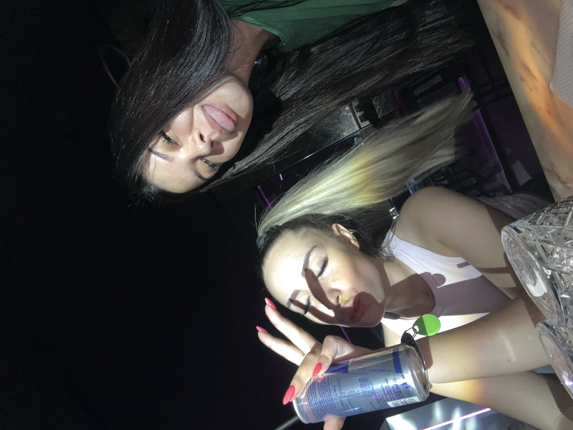 Two young women are pictured at a table in what appears to be a dimly lit nightclub or bar, possibly in Sliven, Bulgaria. The woman on the left, with blonde hair pulled into a ponytail, is holding a red bull can and making a peace sign with her fingers, her eyes are closed and her mouth is pursed. The woman on the right, with long dark hair, is sticking her tongue out at the camera, her eyes are open and she is smiling. They are both wearing casual clothing. The table in front of them has glassware and a white napkin. The background shows a dark, potentially urban, environment with some colored lights.
