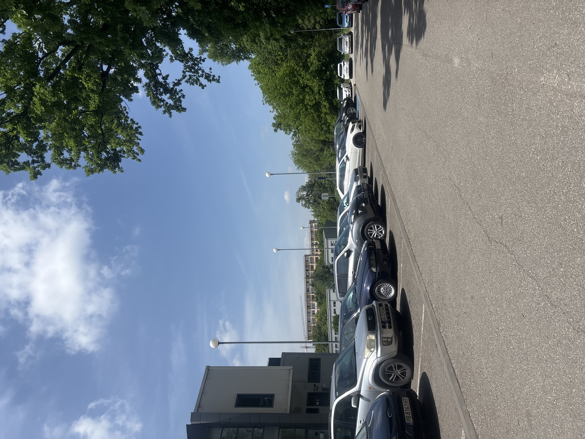 The image shows a row of parked cars along a street in Plovdiv, Bulgaria. The vehicles are parked on the right side of the road. The road surface is asphalt. There are trees along the left side of the road, casting shadows. On the left, there's a building, and in the background, other buildings are visible under a partly cloudy sky. Streetlights are placed along the road. The cars are facing away from the viewer.