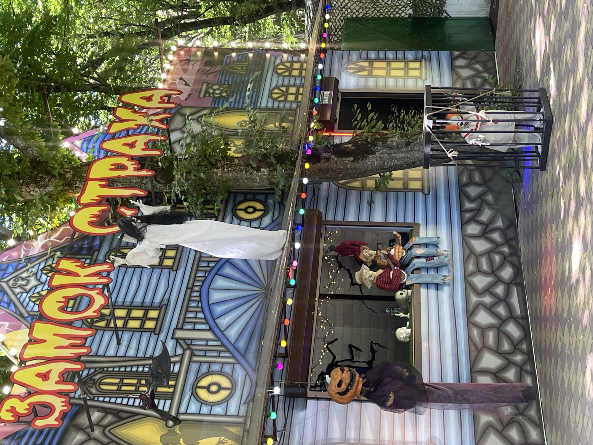 An outdoor Halloween-themed attraction is depicted, likely located in Stavropol, Russia, given the Cyrillic signage. The attraction features a facade painted to resemble a spooky, blue-sided house with yellow-paned windows. Above the structure, a large, yellow, dripping sign in Cyrillic letters reads "ЗАМОК СТРАХА" (Castle of Fear). A white, ghostly figure with long black hair is suspended from the sign, its arms outstretched.
Decorations adorn the facade. To the left, a hanging figure with a jack-o'-lantern head is suspended by a dark cloth. On the ledge of the house's second story, two scarecrow-like figures are seated. One wears a red plaid shirt and a straw hat, and the other is dressed in denim overalls and a straw hat; they appear to be playing instruments. Skulls are placed on the ledge as well.
A string of colorful, illuminated bulbs runs along the roofline above the ledge. To the right, a black cage contains a clown figurine. The clown wears a white outfit with red accents and has a painted face with exaggerated features. The ground in front of the attraction is paved with patterned tiles. Trees with green foliage surround the scene, suggesting an outdoor, possibly park-like, setting. A green, latticed fence is visible on the far right. A sign with the Cyrillic word "ВЫХОД" (Exit) is mounted above a dark doorway on the right side of the building.