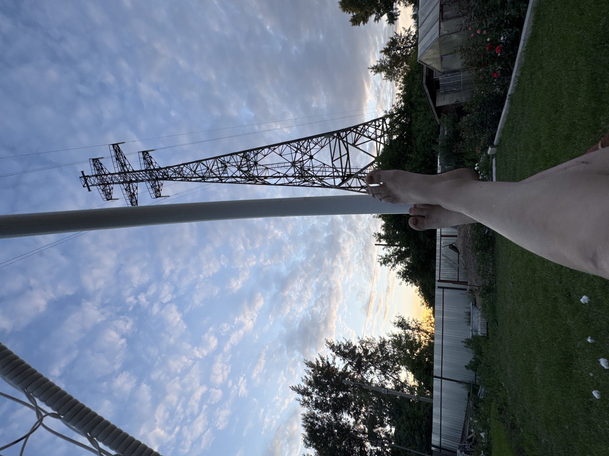 The image shows a person's legs and feet resting in the foreground, with a grassy yard, a fence, trees, a greenhouse, and a large power line tower in the background. The sky is cloudy with hints of sunset colors. The scene appears to be in a rural or suburban area, possibly a backyard. The location is identified as the city of Chaikovsky, Russia. The person's legs are relaxed, suggesting a moment of rest or leisure. The overall atmosphere is peaceful and serene.