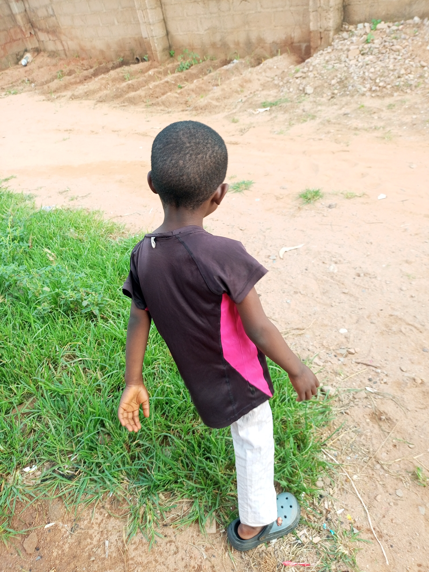 The image shows a young child facing away from the camera, standing on a dirt path in what appears to be an outdoor, undeveloped area. The child is wearing a dark t-shirt with a pink panel on the side and white pants, and a dark clog-style shoe on their right foot. To the left of the child is a patch of green grass. In the background, there is a brick wall and a sloping area covered in dirt and loose stones. Small pieces of debris, such as white paper scraps and colored bits, are scattered on the ground. The overall environment suggests a rural or semi-urban setting.