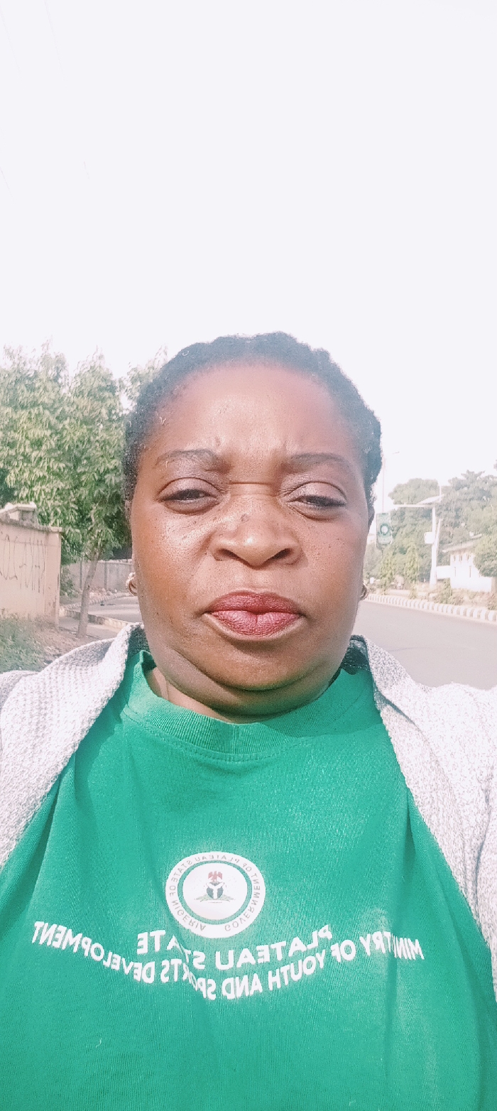 The image captures an adult individual, appearing female, with dark skin, centered and facing the camera. Her facial expression displays a grimace, characterized by furrowed brows, slightly squinted eyes, and tightly pressed, reddish lips. Small hoop earrings are visible in both ears. She wears a bright green t-shirt, partially covered by a light grey, textured cardigan or jacket. The green t-shirt features a circular emblem with the Nigerian Coat of Arms and text, which, when corrected from its mirrored appearance, identifies it as "Plateau State Ministry of Youth and Sports Development." The background is an outdoor urban environment under bright, possibly hazy, daylight. To the left, a light-colored wall with faint markings and green trees are visible. To the right, a paved road extends into the distance, bordered by trees, distant buildings, and a signal pole with what appears to be a dark green traffic light. The scene is located in the city of Kaduna, Nigeria.