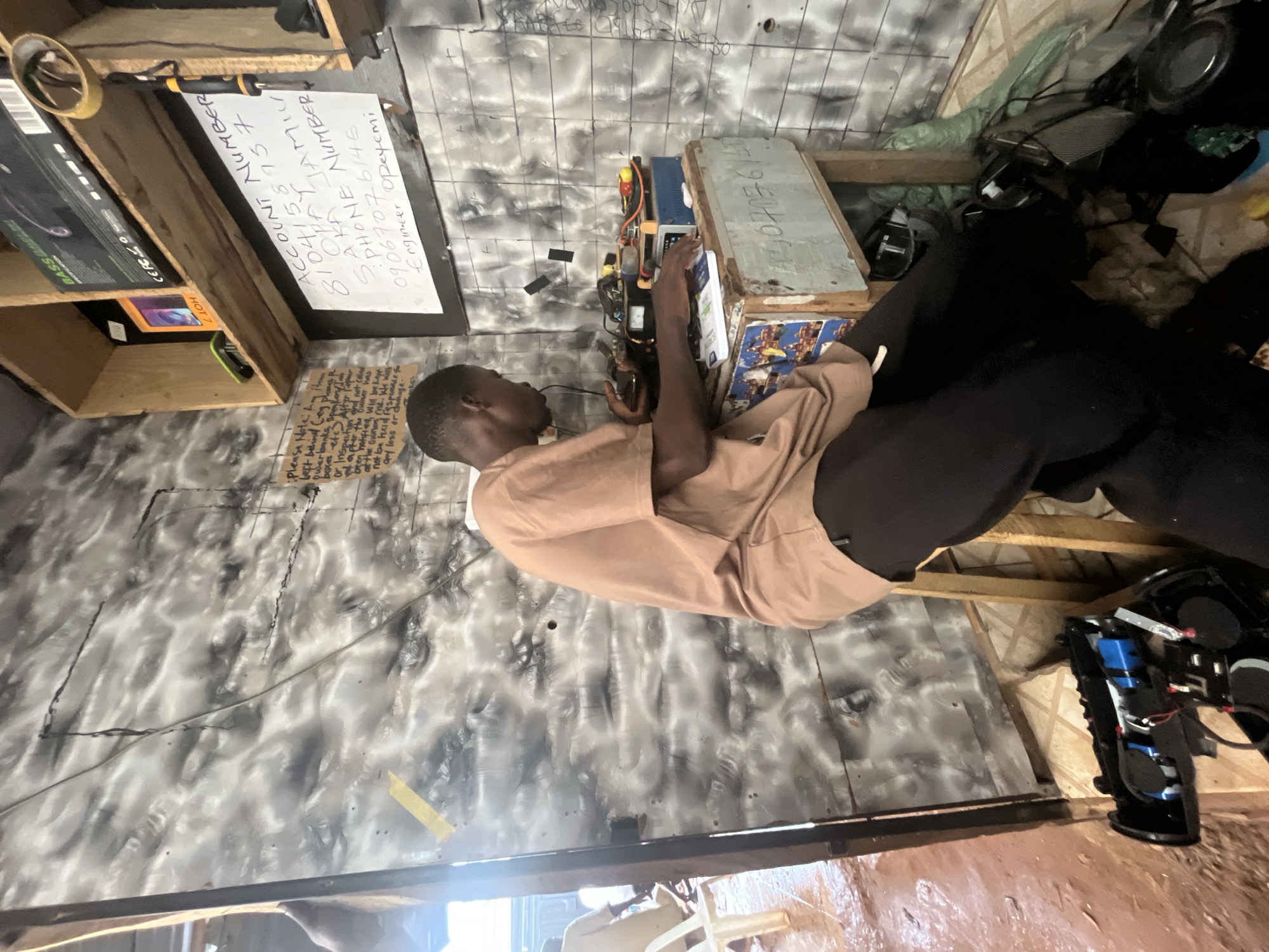 The image depicts a young Black man, likely in his late teens or early twenties, wearing a light brown t-shirt and dark pants, seated at a makeshift workbench. He appears to be engaged in repairing electronic devices. The workbench is cluttered with various electronic components, wires, tools, and a small blue electronic device. Behind him, on a shelf, are packaged electronics, including a "BASS" branded item and another box with "HOT" printed on it. A handwritten note is attached to the wall, listing an account number, a name "SAKA JAMILU," a phone number "09067076145," and "Engineer Opeyemi." Another smaller, handwritten note to the left advises customers to leave phones with the cashier and states the business is not responsible for loss or damage.
The environment appears to be a small, somewhat disorganized workshop or repair shop. The walls are covered in a gray and white mottled material, possibly painted or wallpapered. The floor is a mixture of dirt and possibly water, suggesting an outdoor or semi-outdoor setting, common in areas experiencing rain. In the background, another person in a similar t-shirt is visible, and a white plastic chair is present. Various other electronic items, including a disassembled speaker system, are scattered on the floor around the workbench. The overall impression is of a functional, albeit humble, electronics repair business located in a less developed urban setting, consistent with Ado, Nigeria.