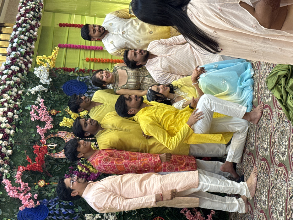 In Malasar, India, an indoor scene depicts a group of ten individuals, predominantly male, posing for a photograph. The background features a green wall extensively decorated with artificial flowers in shades of pink, white, red, and yellow. Two circular blue woven ornaments and a central gold-colored crescent moon and sun motif are affixed to this floral wall. To the right, a light green fabric curtain is visible, adorned with vertical garlands of red flowers. The floor is covered by a patterned carpet with a green base and intricate maroon designs.

Nine individuals are arranged in a multi-level pose. Six men are standing: one in a light peach kurta and white churidar; one in an orange and red patterned kurta and white churidar; two in bright yellow kurtas, one with white churidar; one in a pale yellow kurta with dark sunglasses; and one in a white and gold patterned kurta. One woman stands in a green patterned choli and skirt. In the foreground, one man kneels on his right knee, wearing a bright yellow kurta and white churidar. Adjacent to him, a woman sits wearing a yellow and light blue ombre saree/lehenga with a white top; intricate henna patterns are visible on her hands. Another man sits nearby in a light peach patterned kurta. All visible faces display smiles, and most are directed slightly to the right of the camera's perspective. Several men are barefoot. In the immediate foreground, the back of a third woman, wearing a light peach top, is partially visible, facing away from the camera, with henna on her right hand. A folded piece of green fabric rests on the carpet in the lower foreground.