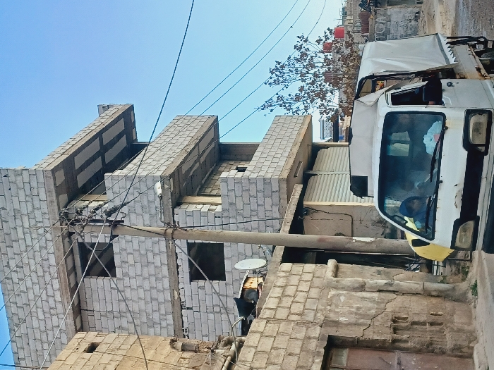 The image depicts an urban street scene in Kafr Sousa Municipality, Syria, under a clear blue sky. A white, visibly aged pickup truck with a white canvas bed cover occupies the lower right and center-front. Two individuals are seated within the truck's cab; the passenger, on the left, wears a blue head covering.
A concrete utility pole stands centrally, laden with numerous black electrical or communication wires and multiple cross-arms. To the left of the pole, an older, multi-story building constructed of light-colored concrete blocks exhibits signs of wear and cracking. A satellite dish is affixed to its roof, and a weathered wooden door is visible at ground level.
To the right of the utility pole, a multi-story building, likely unfinished or under construction, is visible. It is constructed from light-colored concrete blocks, featuring exposed concrete floor slabs serving as balconies or overhangs. Window openings are present but lack frames or glass. The ground level of this structure displays two storefronts with white corrugated metal roll-down shutters.
In the background, behind the truck and further right, a leafless tree and additional residential structures with red rooftop water tanks are discernible. The overall environment suggests a densely built-up area.