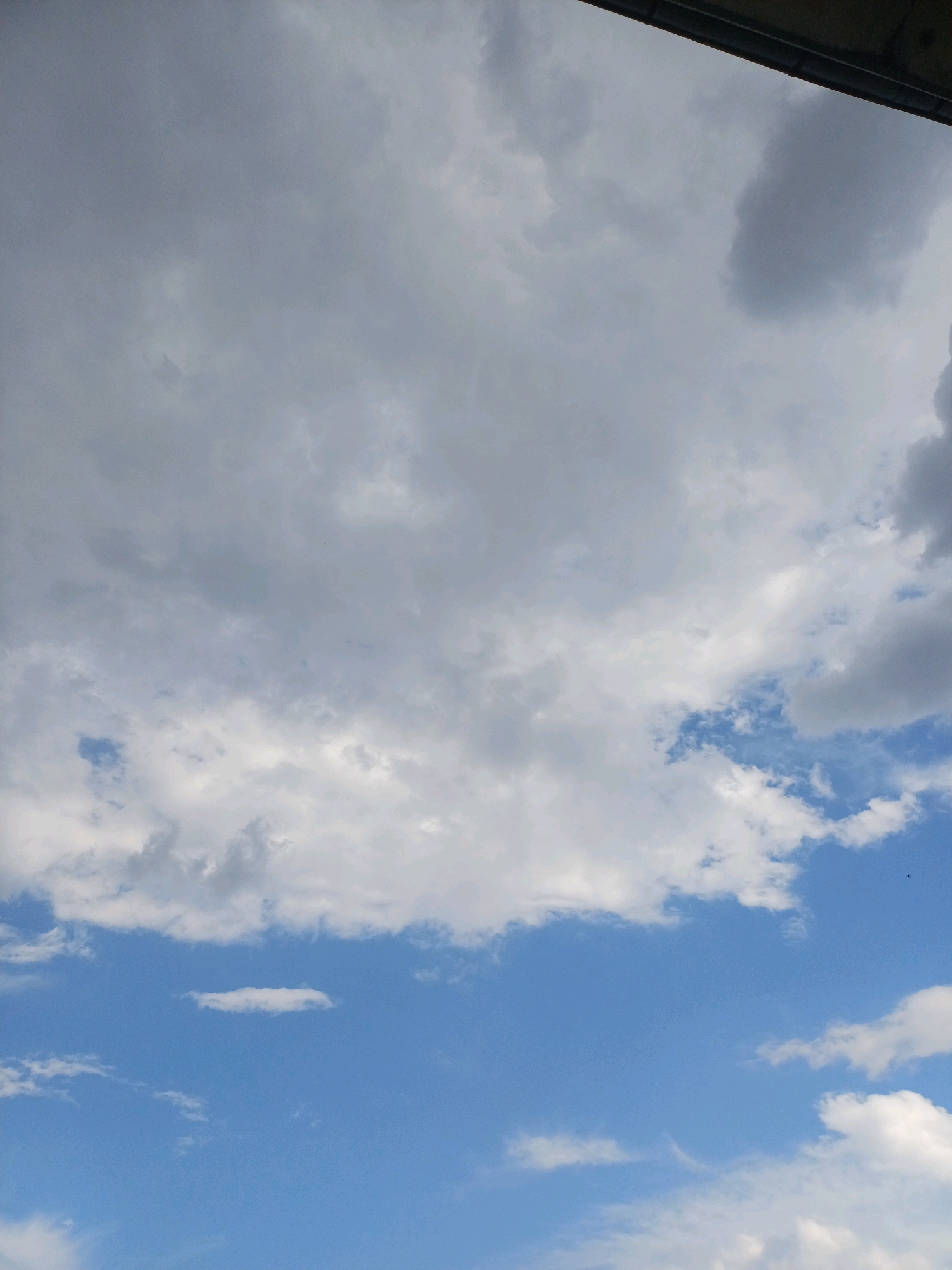 The image depicts a sky filled with a mixture of clouds over Cheshnegirovo, Bulgaria. A dark, horizontal structure, likely an overhang or building edge, is visible at the top of the frame. The clouds range from dense, gray masses in the upper portion to lighter, white formations and patches of clear blue sky in the lower portion. The scene is static, with no people or discernible action. The environment is characterized by the sky and its cloud cover.