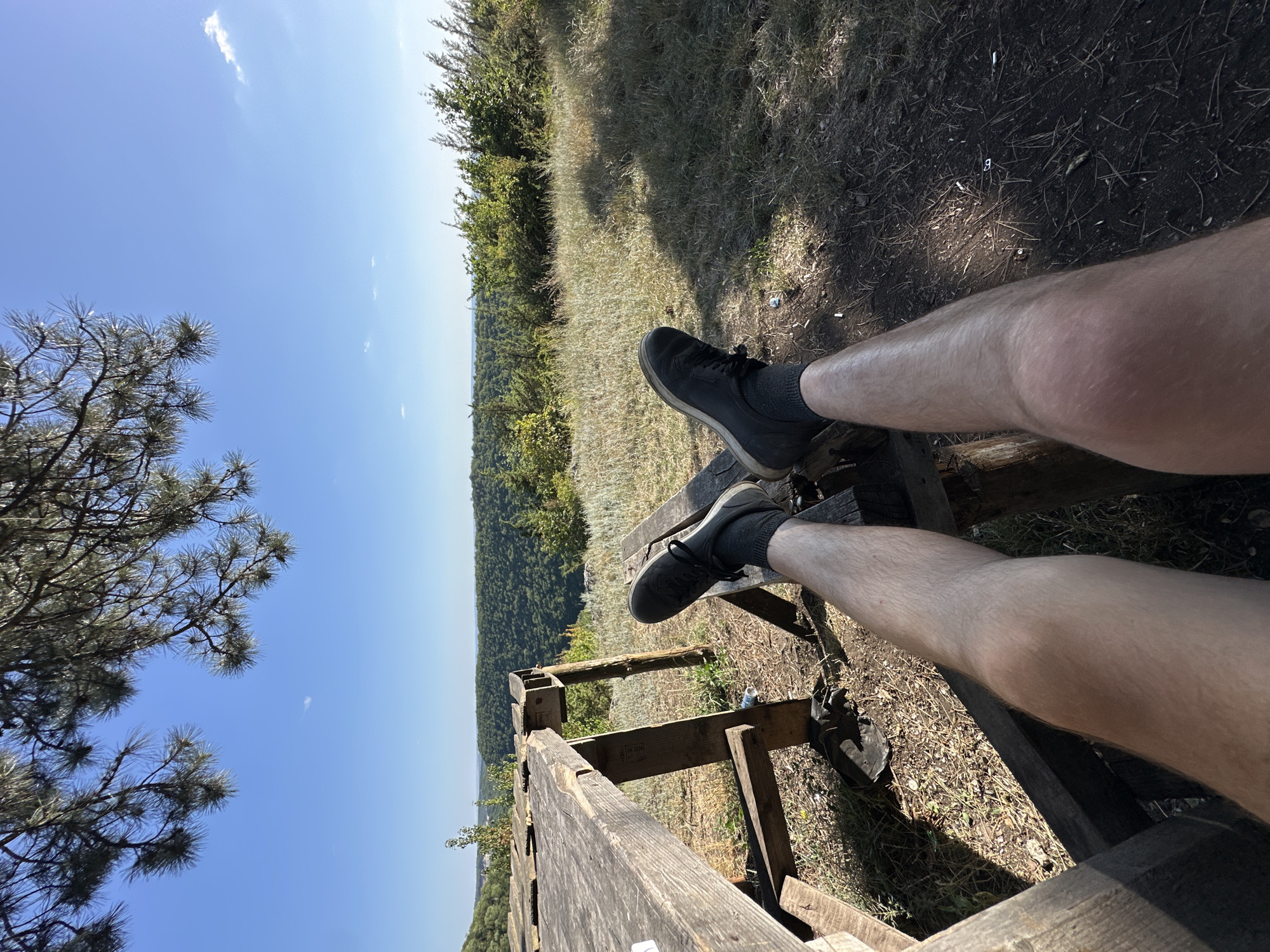A first-person perspective shot shows a person's legs and feet resting on a wooden bench. The person is wearing dark sneakers and dark socks. The background depicts a scenic overlook with a vast expanse of green forest and hills under a clear blue sky with sparse clouds. The sun is shining brightly, indicating it is daytime, likely midday or afternoon. The scene appears to be in a natural, outdoor setting, possibly a park or hiking trail. The overall mood seems relaxed and peaceful, with the person enjoying the view. There is a wooden railing in the foreground, and the ground is covered with dry grass and dirt. The location is Malozalissia, Ukraine.