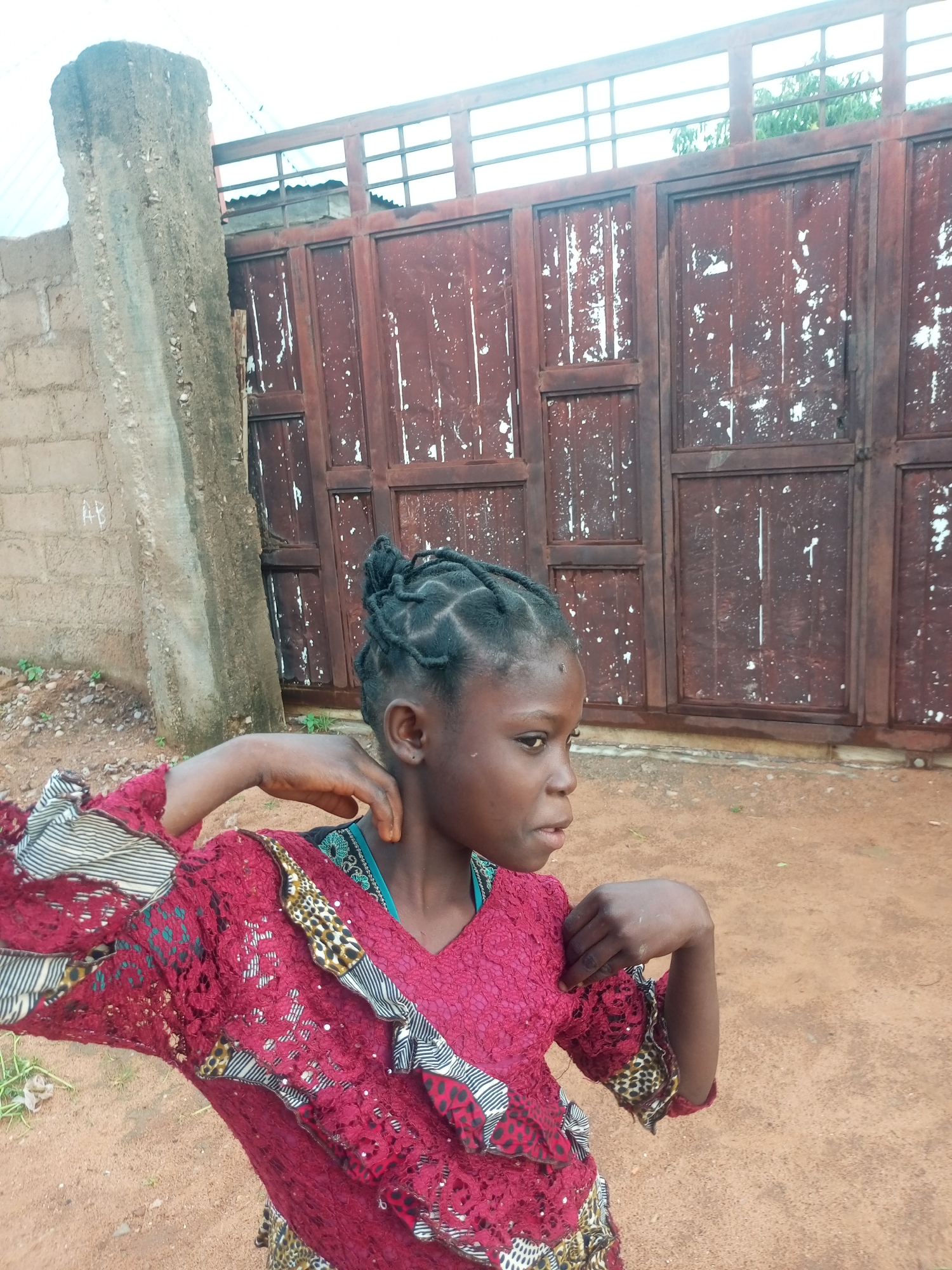 A young girl with braided hair stands in front of a rusty, double-gated entrance, likely in Tempure, Nigeria. The girl is wearing a red lace dress with colorful patterned sleeves. Her right arm is raised, and she appears to be adjusting her dress or hair. The background consists of a rough-textured concrete pillar and a brick wall to the left, and the weathered metal gates with peeling paint to the right. The ground is sandy and reddish-brown, with some sparse vegetation. The overall scene suggests an outdoor setting in a rural or semi-urban area.