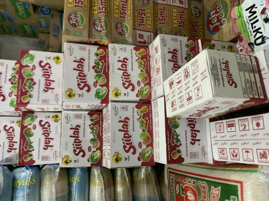 The image shows a stack of boxes of "Siplah" fruit juice in a shop in Kecamatan Lawang, Indonesia. The boxes are white with red text and feature images of various fruits. Some boxes are stacked on top of other boxes, and others are arranged in a single row. The boxes are all sealed. There are also other boxes of different brands visible behind the "Siplah" boxes. A few blue and white items are visible in the lower part of the image, but their details are unclear. The background is a wall with some printed graphics.