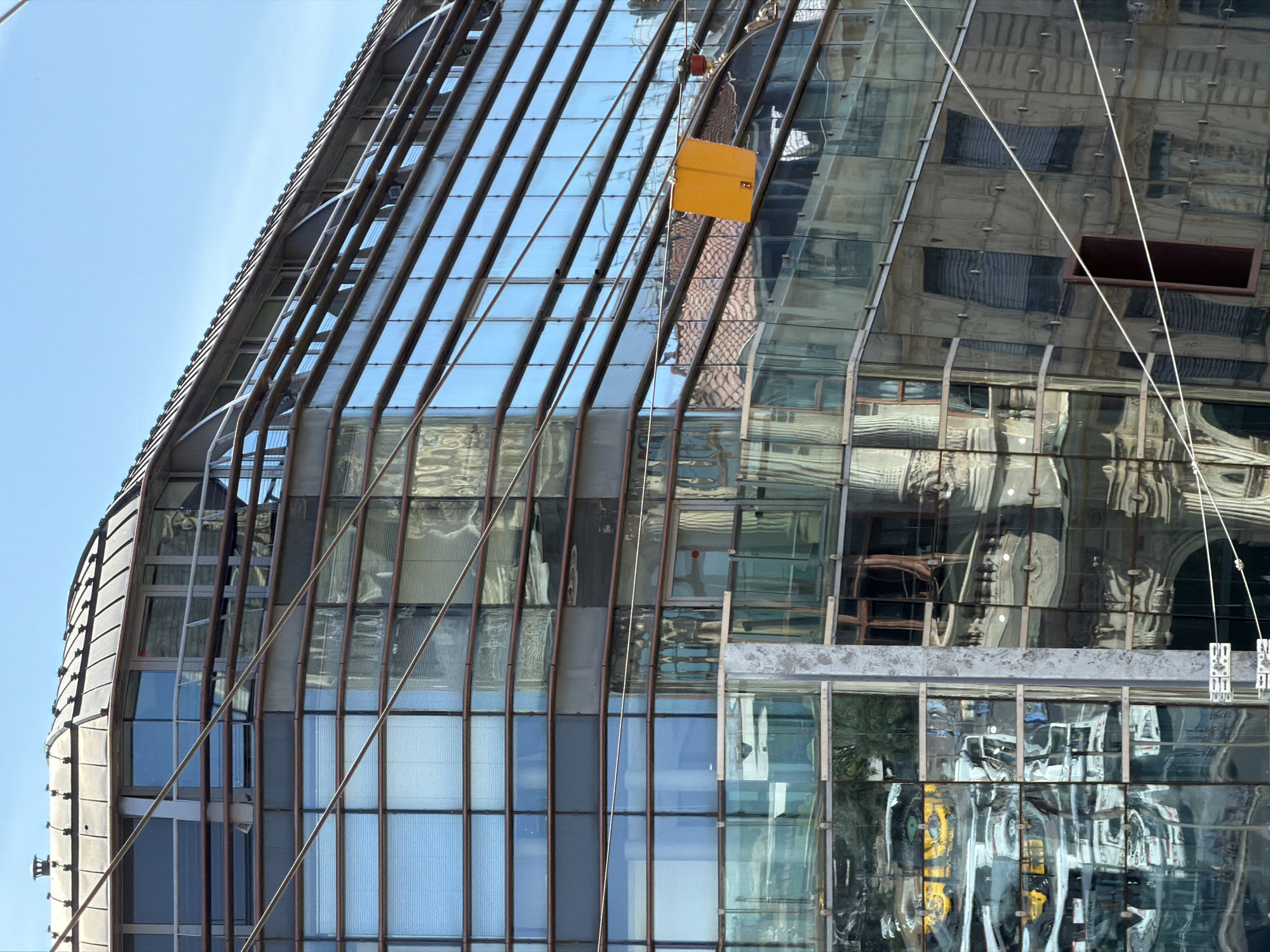 This image captures a modern glass-fronted building with reflections of older architecture. The building has a curved roof and a multi-paneled glass facade. Horizontal metal beams are interspersed throughout the glass. In the reflection, an ornate, classical building with arched windows and shutters is visible. On one section of the glass facade, a bright orange rectangular sign is attached to a wire. The reflection also shows a bright yellow car and white vehicles, suggesting a street or parking area. The sky above is clear and blue. This building is located at 77 Rue de la République in Lyon, France.