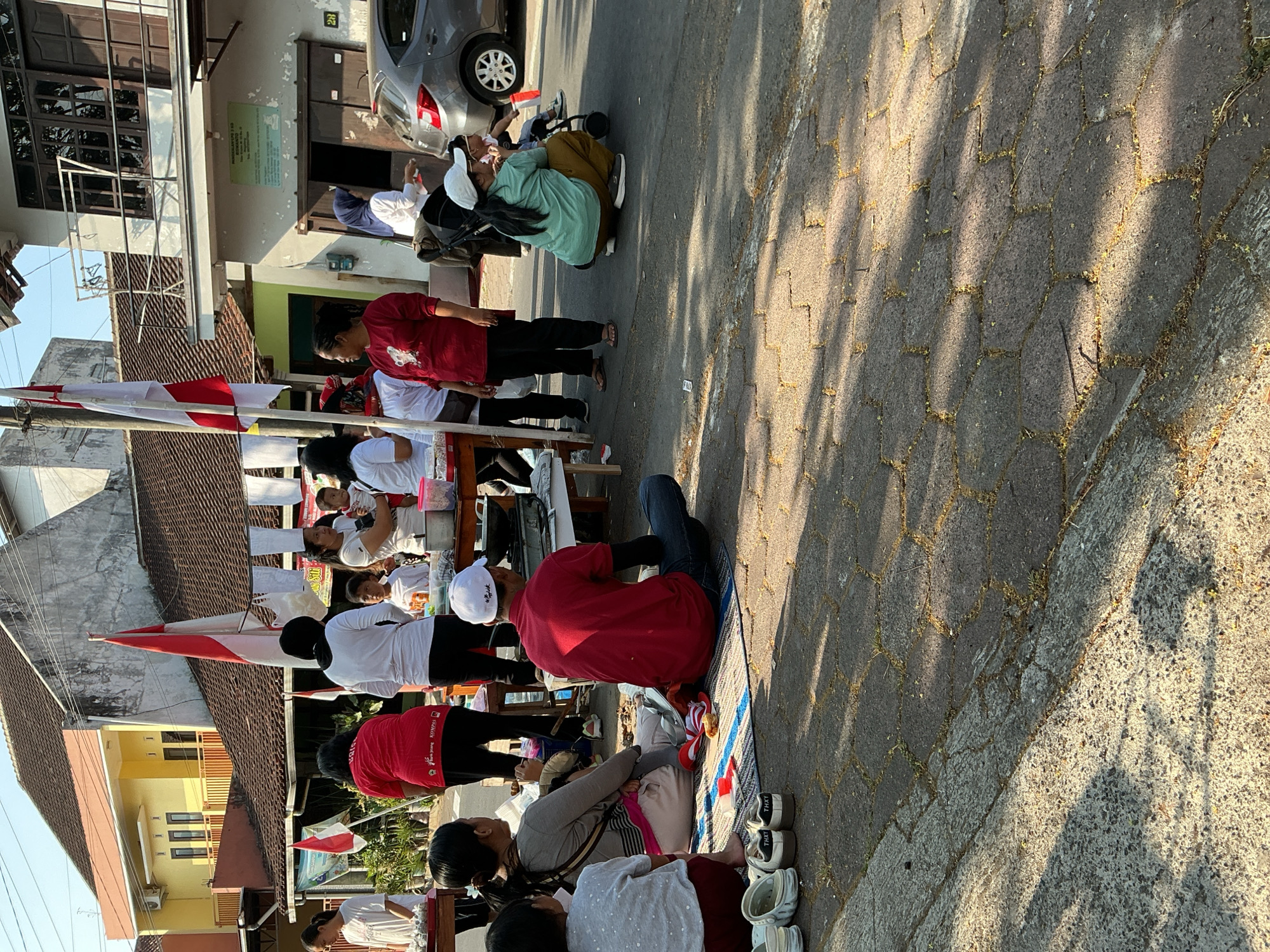 In Magelang, Indonesia, on a street with patterned brick paving, a gathering of people is observed outdoors. The scene is illuminated by natural sunlight, casting distinct shadows. On the left, several individuals, including two children and an adult woman in a red shirt, are seated on a blue and white patterned mat. The woman is holding a baby. In the center, a man in a red shirt and dark pants is sitting on the mat, facing away from the camera. To his right, a table with food items and cooking utensils is visible. In the background, a group of people are standing, some holding flags, including one that is red and white. Buildings with tiled roofs and windows line the street, suggesting a residential or commercial area. A grey car is parked on the right side of the image, and a stroller is also present. The overall atmosphere appears to be a casual outdoor event or market.