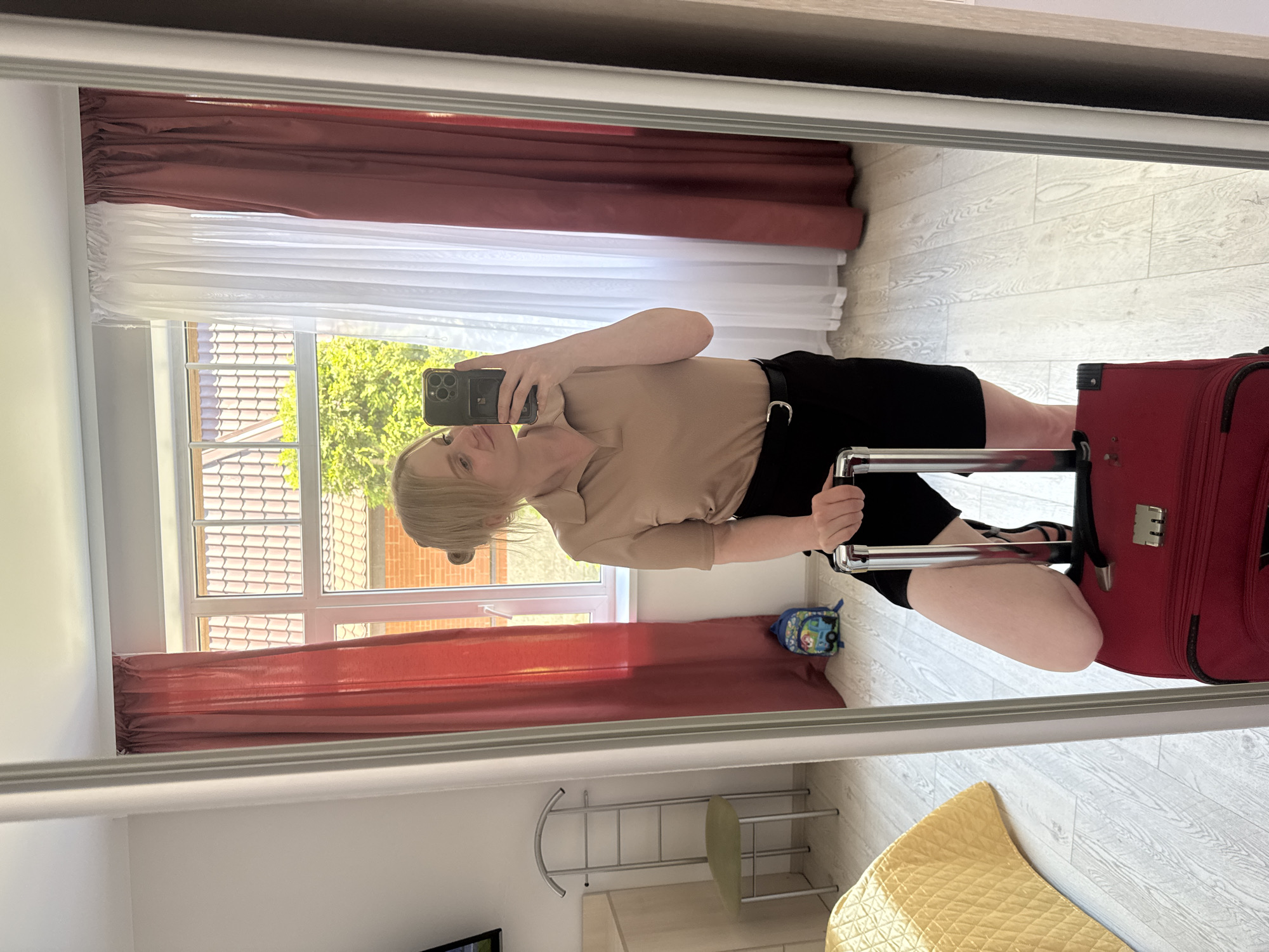 The image shows a mirror reflection of a woman holding a smartphone and taking a selfie. She is wearing a beige collared shirt and black shorts with a black belt. She is kneeling next to a red suitcase. The scene appears to be indoors, possibly a hotel room, with a window in the background showing trees and tiled rooftops, suggesting a daytime setting. The room has wooden floors and is decorated with red and white curtains. A small backpack with cartoon characters is visible on the floor. The reflection also captures a metal garment rack and a yellow quilted cushion. The woman's expression is neutral. There is no visible text in the image.