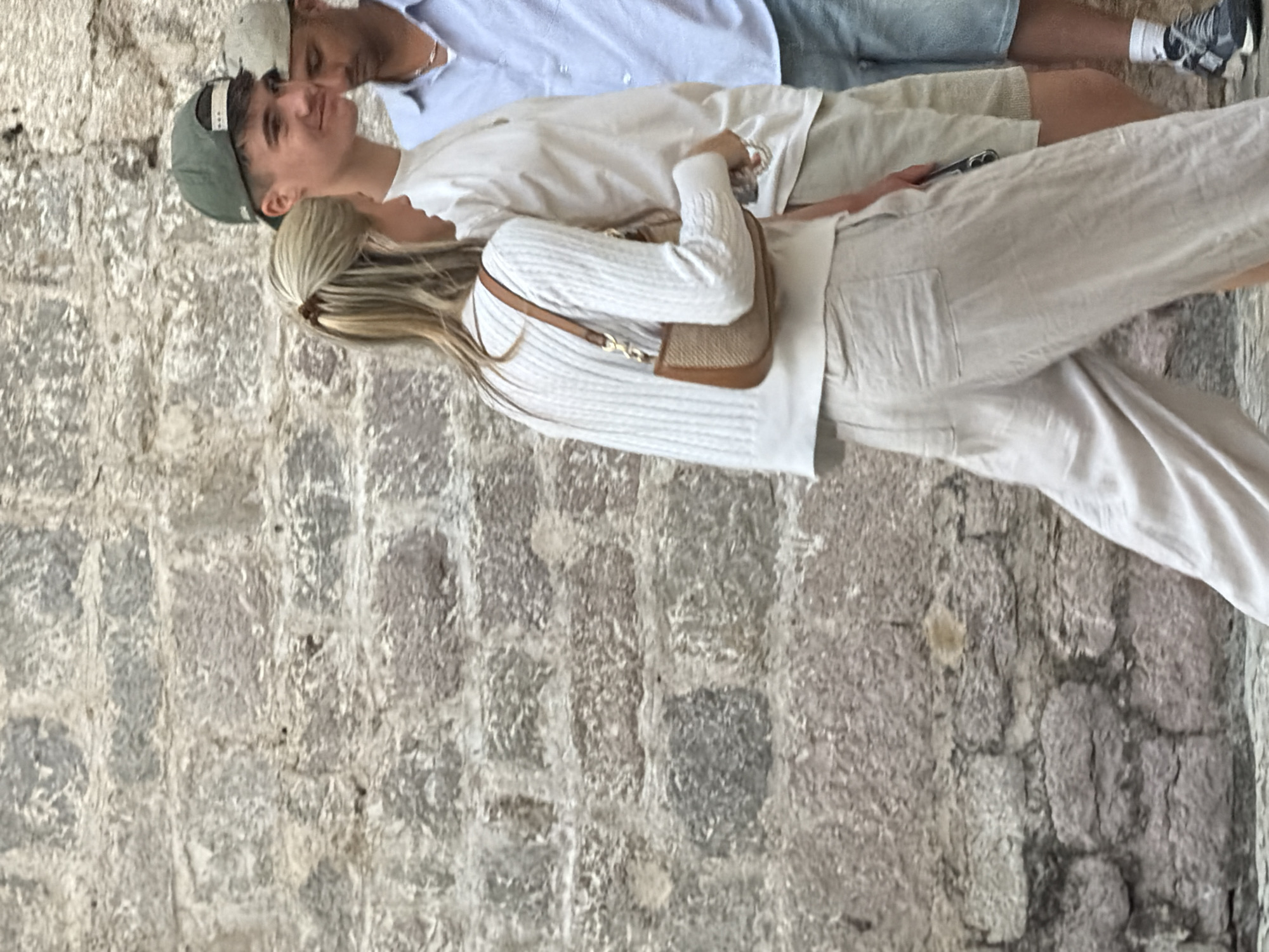 In Budva, Montenegro, three individuals are captured in a candid moment against a textured stone wall. A woman in the foreground, with blonde hair pulled back, wears a white cable-knit sweater and loose, light-colored linen pants. She carries a small, tan shoulder bag. To her right, a young man sporting a green baseball cap and a white t-shirt, smiles as he looks off to the side. Partially visible behind him is another man wearing a light blue collared shirt and a light-colored cap, also looking away. The scene is likely a street in Budva's Old Town, characterized by its ancient stone architecture.