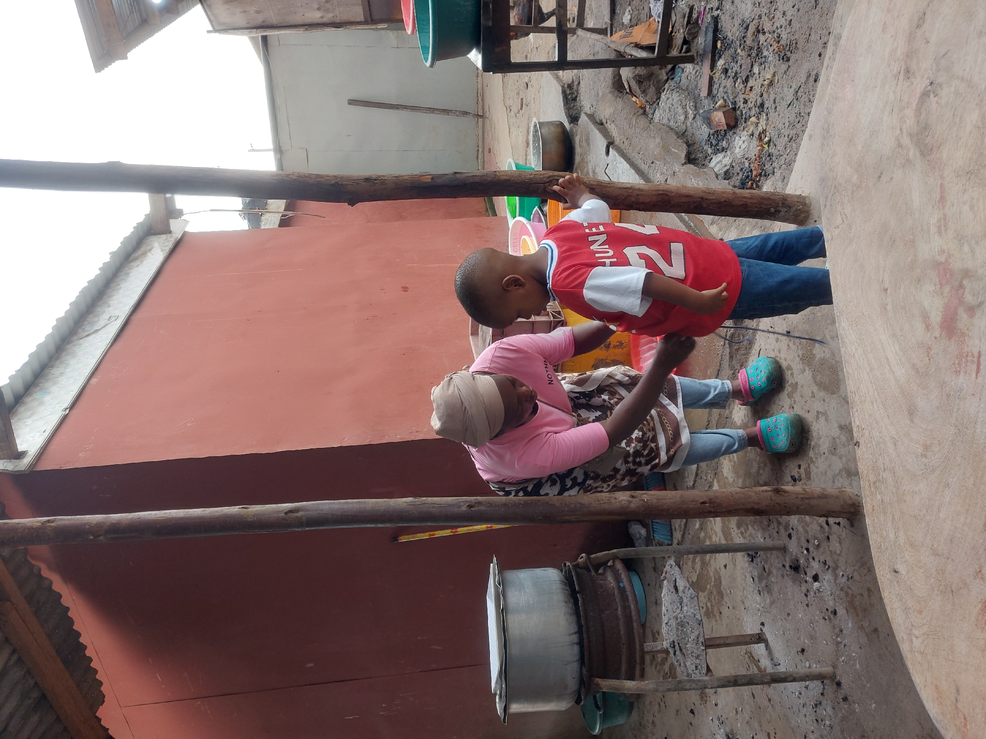 The image shows a child in a red shirt with the number 22, and blue jeans, next to a woman wearing a pink shirt, a patterned skirt, and a head covering. They are both standing next to a wooden pole that seems to form part of a makeshift structure. Beside the woman is a large metal pot on a rudimentary stove. The ground appears to be a dirt surface with scattered debris and ash. In the background, there is a red building and other structures indicating a densely populated area, potentially suggesting an urban setting in Dar es-Salaam, Tanzania. The interaction between the child and woman suggests a familial connection.