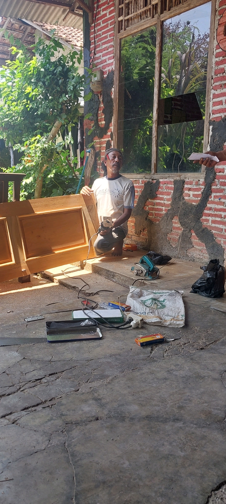 A man is kneeling on a cracked concrete floor in front of a partially constructed wooden door. He is wearing a light-colored t-shirt, dark pants, and a cap. In his right hand, he holds a woodworking tool, possibly a router. To his left, the wooden door is propped up, revealing its unfinished surface. The scene appears to be an outdoor workshop or yard, with a brick wall and a large window in the background. Various tools and materials, including a saw, a planer, a bag, and electrical cords, are scattered on the ground. Lush green foliage and trees are visible behind the man and to the left of the structure, suggesting a tropical or subtropical environment. The setting suggests a carpentry project is underway. The location is Keling, Jepara, Indonesia.