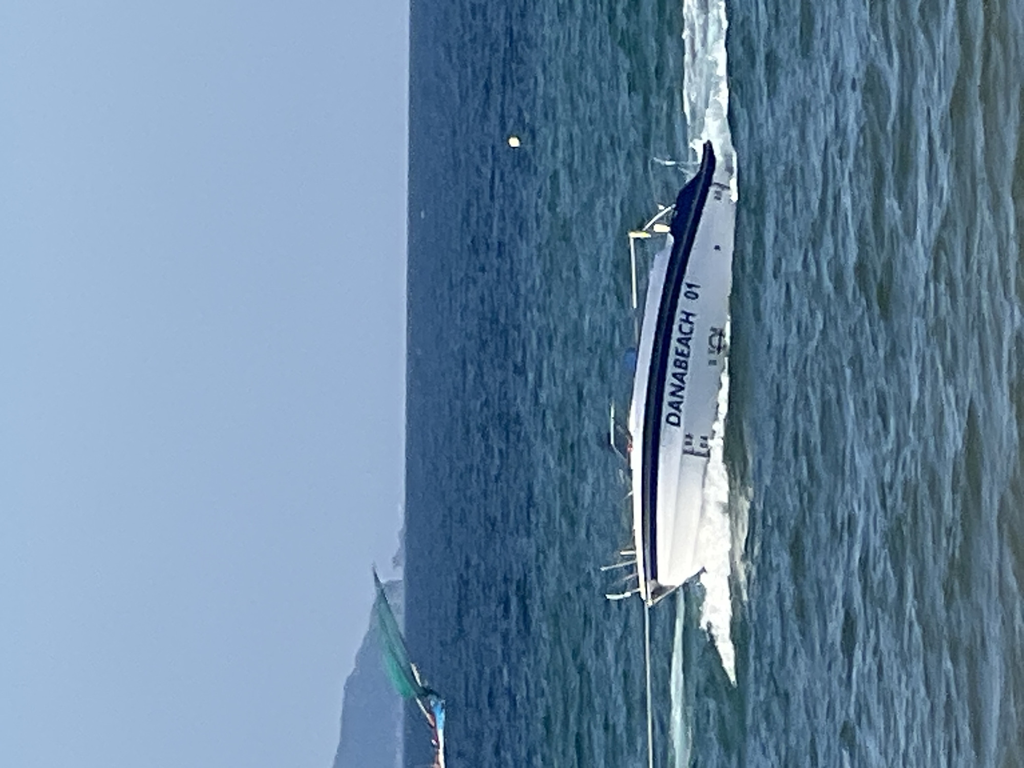 A white speed boat with "DANABEACH 01" printed on its side is moving through blue water, leaving a white wake. The boat has a dark blue stripe along its upper edge and a metal rail on top. In the background, there is a vast expanse of blue ocean under a clear, light blue sky. To the left, partially visible, is another boat with a green sail or canopy. In the far distance, a hazy, mountainous coastline is visible. A small yellow buoy floats on the water to the right of the speed boat. The scene is set on a bright, sunny day, likely in the waters off Đà Nẵng, Vietnam, given the boat's labeling.