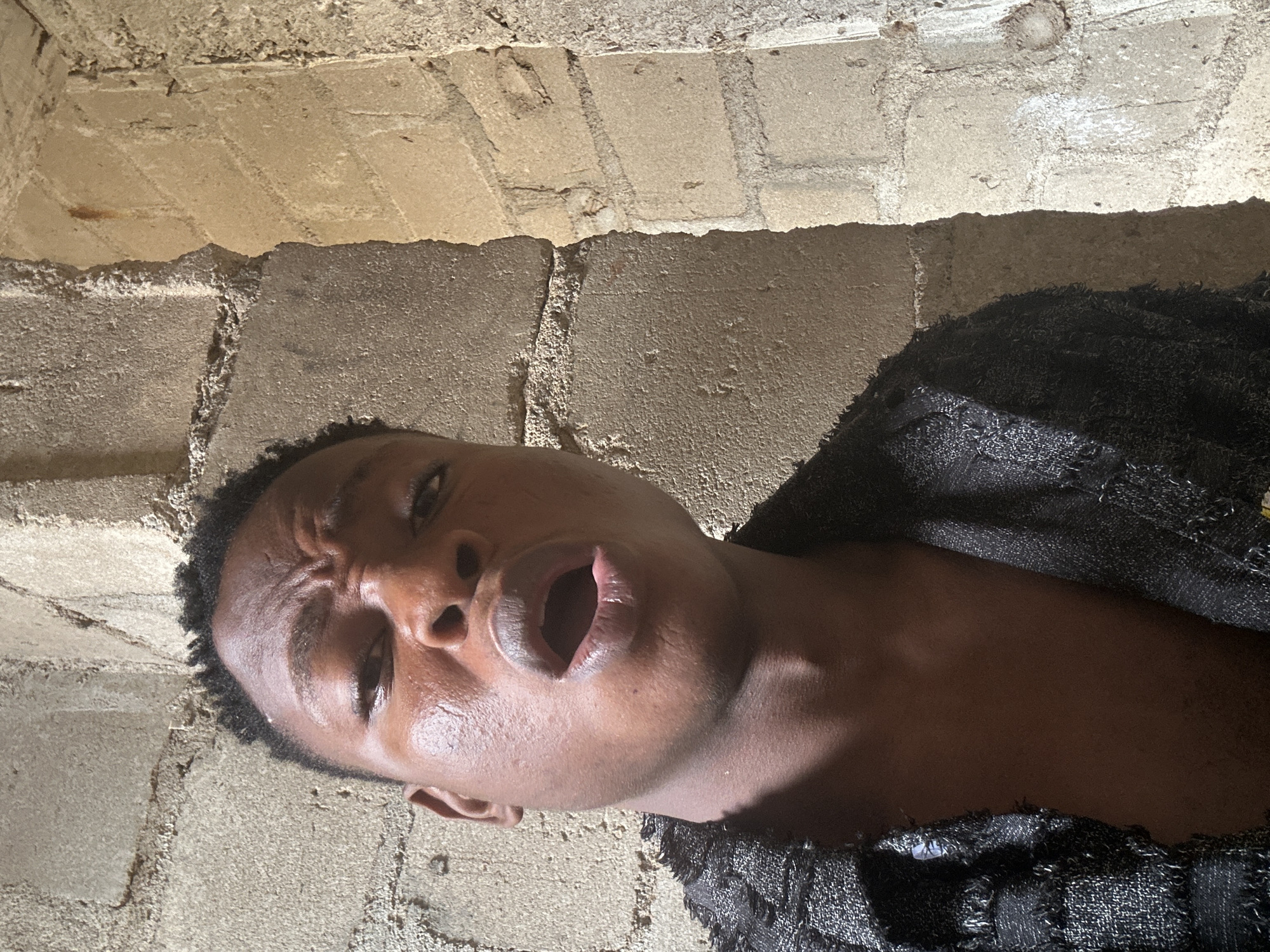 A young Black male is captured in a close-up, low-angle selfie. He has dark, short hair and his face is angled towards the camera, with his mouth open. He is wearing a black garment with a textured, woven appearance and fringed edges. The background consists of textured concrete walls, suggesting an urban or industrial setting. The lighting appears to be natural and somewhat harsh, casting shadows on his face and the walls. The image does not contain any visual cues to specifically place it within Ikorodu, Nigeria.