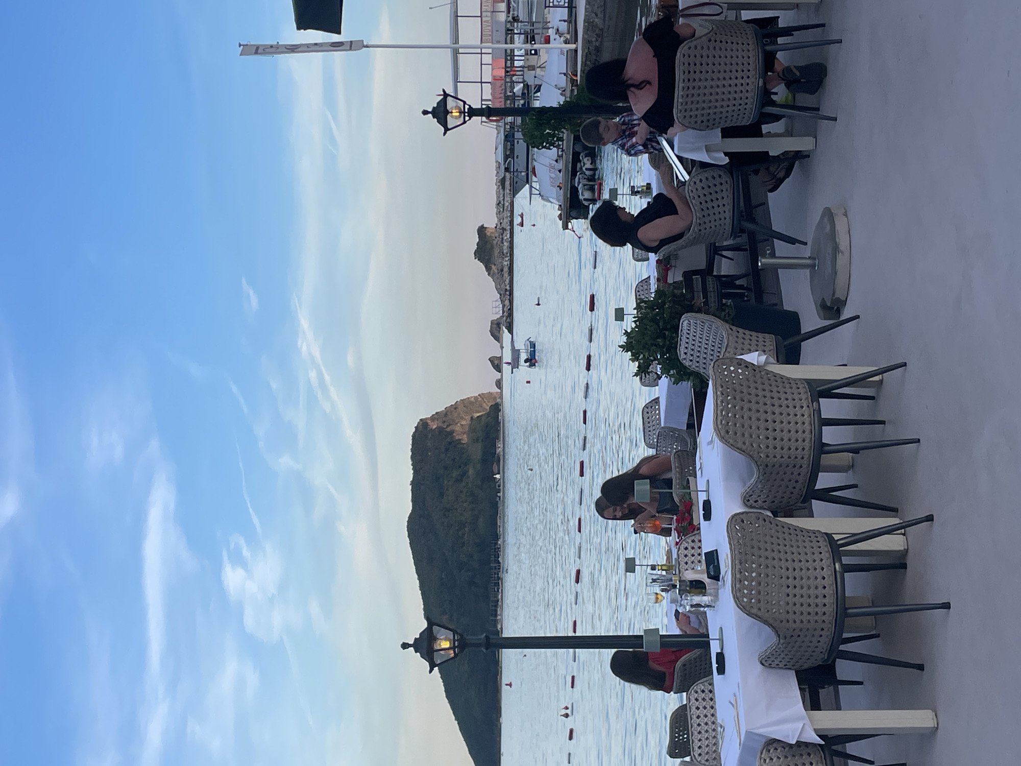 The image depicts an outdoor seating area of a restaurant or cafe in Budva, Montenegro, overlooking the Adriatic Sea. In the foreground, several tables are set with white tablecloths and chairs. People are seated at some of these tables, engaged in dining and conversation. One table has a woman in a black dress and a young boy in a plaid shirt. Another table has three women, one in a red top and two in dark attire. A street lamp stands prominently in the left mid-ground.
In the background, the calm sea is visible, with a large, green, mountainous island dominating the horizon. Several boats are moored or moving in the water. The sky is a clear blue with scattered, wispy clouds, suggesting it is late afternoon or early evening. The overall scene conveys a relaxed, social atmosphere.