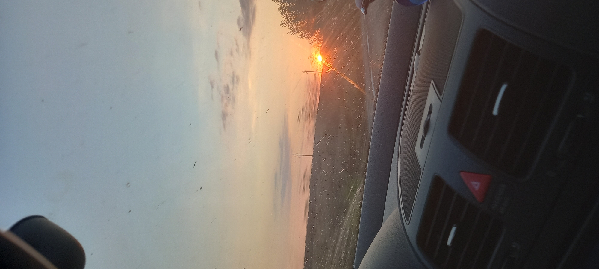 The image is a perspective from inside a car, looking out the windshield at a sunset. The sky transitions from a pale blue at the top to hues of orange and pink near the horizon. Wispy clouds are visible, catching the warm light of the setting sun. The sun itself is a bright orb, partially obscured by a treeline on the right side of the frame and a distant hill on the left. A single utility pole stands on the left side of the road in the middle ground.

The car's dashboard is visible in the foreground, a dark, textured surface with air vents and a hazard light button. The windshield has several smudges and streaks, some of which are highlighted by the light. The view suggests the car is traveling along a road that borders a grassy or field-like landscape.

While the image depicts a scenic sunset, there is no specific indication of the location within Smolnitsa, Bulgaria, other than the potential context provided. The scene is static and captures a moment in time during travel.