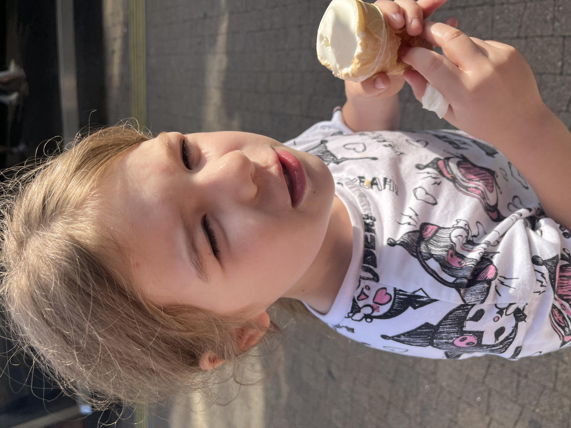 The media file shows a close-up, rotated image of a young girl outdoors, holding an ice cream cone. The girl, with blonde hair, is wearing a white t-shirt with a cartoon print of cat-like characters in black and pink. Her eyes are closed, and her mouth is slightly open as she holds the ice cream. Her hands are visible, one holding the cone and the other holding a small white piece of tissue. The background appears to be a paved surface, possibly a sidewalk or pathway. The lighting suggests it is daytime, with sunlight casting shadows. The girl's expression appears content or perhaps experiencing the cold of the ice cream. The overall scene evokes a casual, summery moment. The location context is Sochi, Russia. There is no visible text in the image.