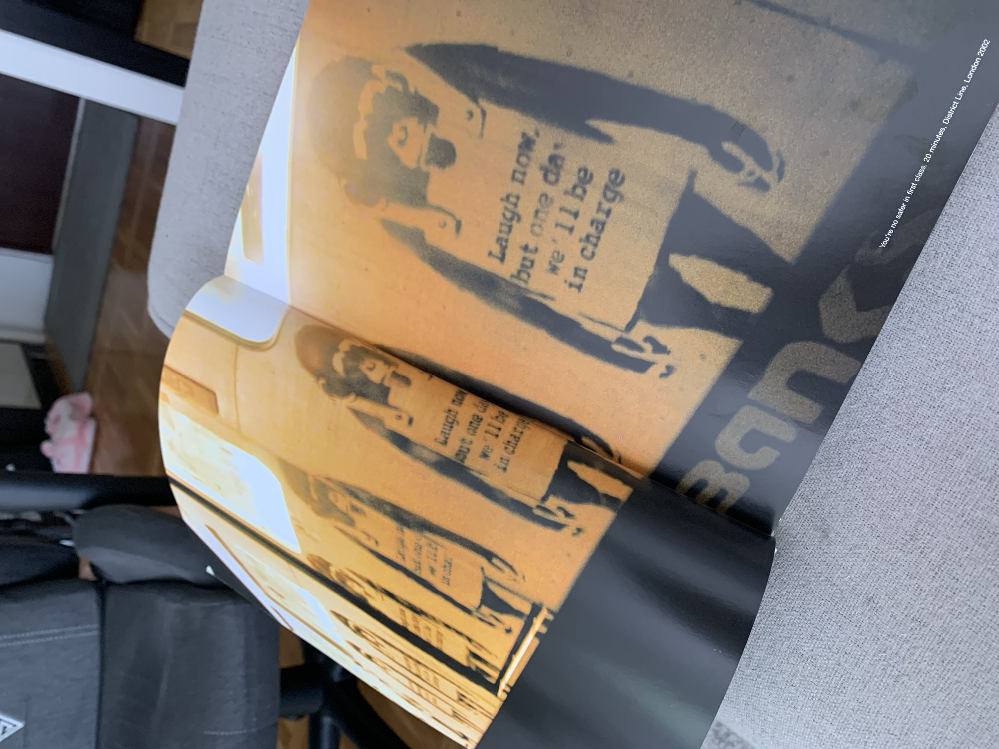 An open book or magazine is depicted at an angle, displaying two pages featuring stencil artwork. The primary artwork, visible on the right page, shows a black silhouette of a figure, possibly a child, holding a sign against an orange-brown background. The sign contains the text: "Laugh now, but one day we'll be in charge." A similar, partially visible stencil of another figure with the same text on a sign is seen on the left page.
Along the bottom right edge of the page, white text provides context for the artwork: "You're no safer in first class. 20 minutes, District Line, London 2002." Part of the artist's name, "Bane" (likely referring to Banksy), is also visible on the right page.
The book's pages have a glossy finish, reflecting light. The setting appears to be indoors, with a light gray upholstered surface visible to the right of the book, possibly part of a chair or sofa. Darker furniture or objects and a glimpse of a wooden floor are visible in the background on the left. The image is a static shot of the open publication.