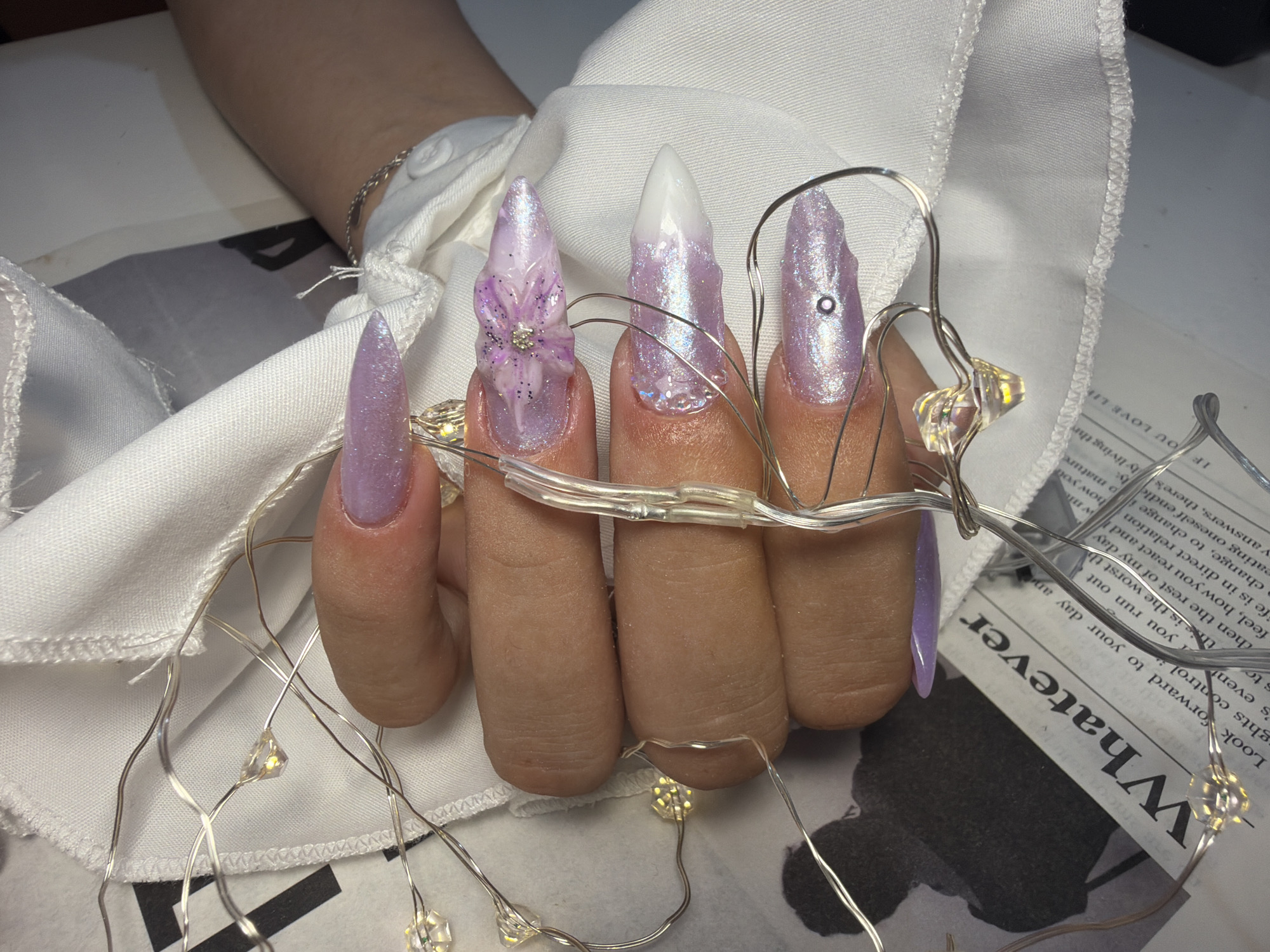 The image captures a close-up view of a right hand, positioned in the city of Stara Zagora, Bulgaria. The hand is adorned with five elongated, pointed acrylic nails, predominantly painted with an iridescent, shimmering light purple polish. The nail on the ring finger features a prominent three-dimensional floral design in a lighter purple shade, detailed with glitter and small rhinestones at its center. The middle finger's nail transitions from a solid white tip to the shimmering purple base. The index finger's nail includes a small, dark circular piercing near its distal end. A delicate silver chain bracelet is visible on the wrist. A strand of fairy lights, characterized by small, clear, faceted LED bulbs emitting a warm glow, is intricately wrapped around and draped over the fingers and hand. The hand rests upon a surface partially covered by a white fabric, possibly a garment or tablecloth, exhibiting a visible stitched hemline. Beneath the white fabric, a section of printed material, resembling a newspaper or document, is partially visible, displaying the word "Whatever" in a large, upside-down serif font, along with smaller, illegible text.