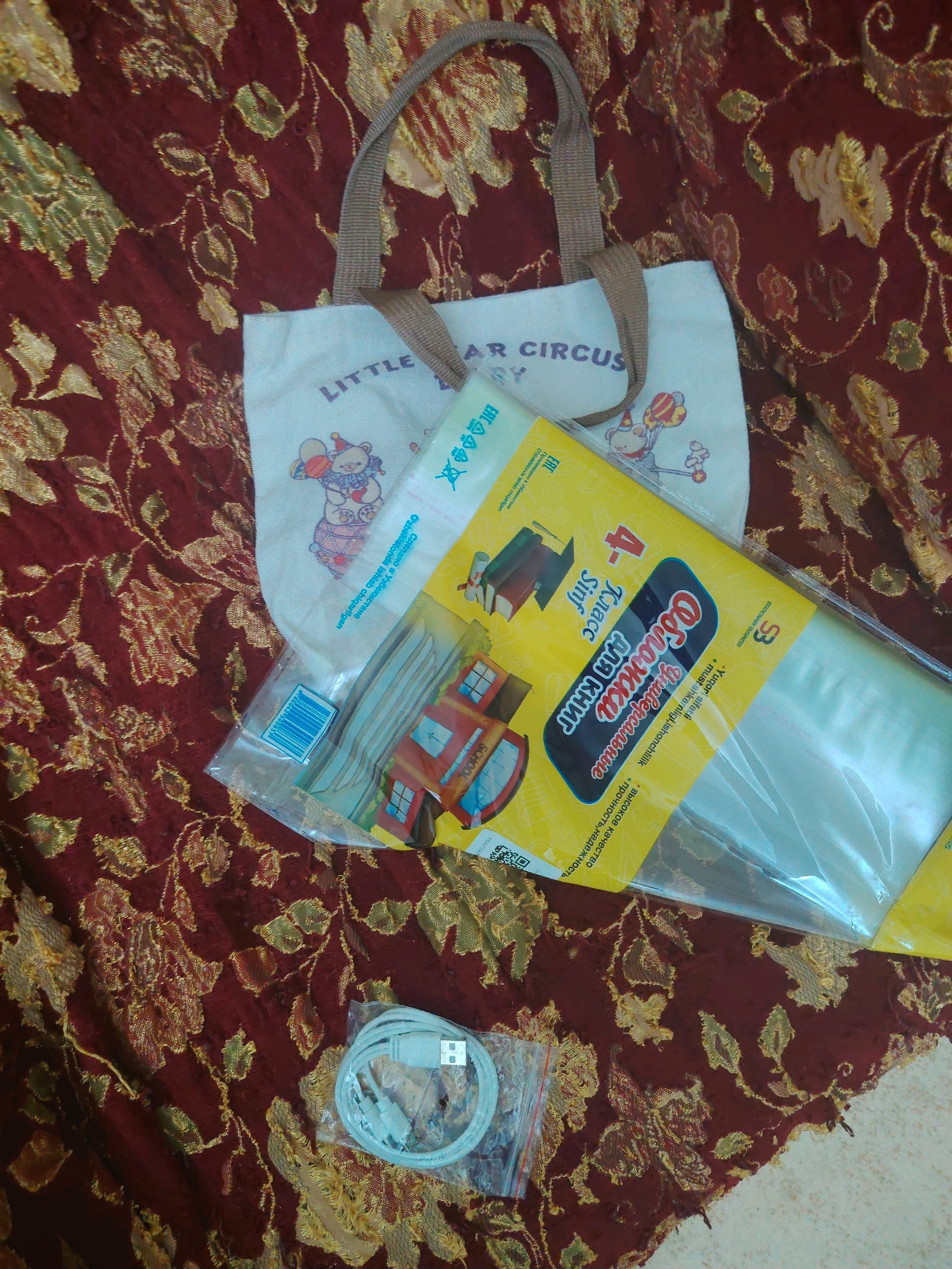 The image depicts a still life arrangement on a textured, dark red fabric with gold floral patterns. The objects are laid out on the fabric, suggesting an indoor setting, possibly a home or a shop.
In the center-left of the image, there is a light-colored canvas tote bag with a woven handle. The bag has a drawing of a teddy bear wearing a hat and holding balloons. The words "LITTLE CIRCUS" are printed in purple lettering above the drawing.
To the right of the tote bag, a clear plastic package contains school supplies. The packaging is predominantly yellow and features cartoon images of a school bus and a school building. Text in Russian is visible, including "4 Класс" (4th Grade) and "Обложка для книг" (Book Cover). There are also some plastic cutlery items visible inside this package.
In the bottom-left corner, a small clear plastic bag contains a coiled white USB cable. The cable is neatly wrapped and appears to be new.
The overall scene suggests a collection of new items, possibly for school or personal use, presented against a decorative textile background. There is no indication of the city of Zaamin, Uzbekistan, within the image itself or its contents.