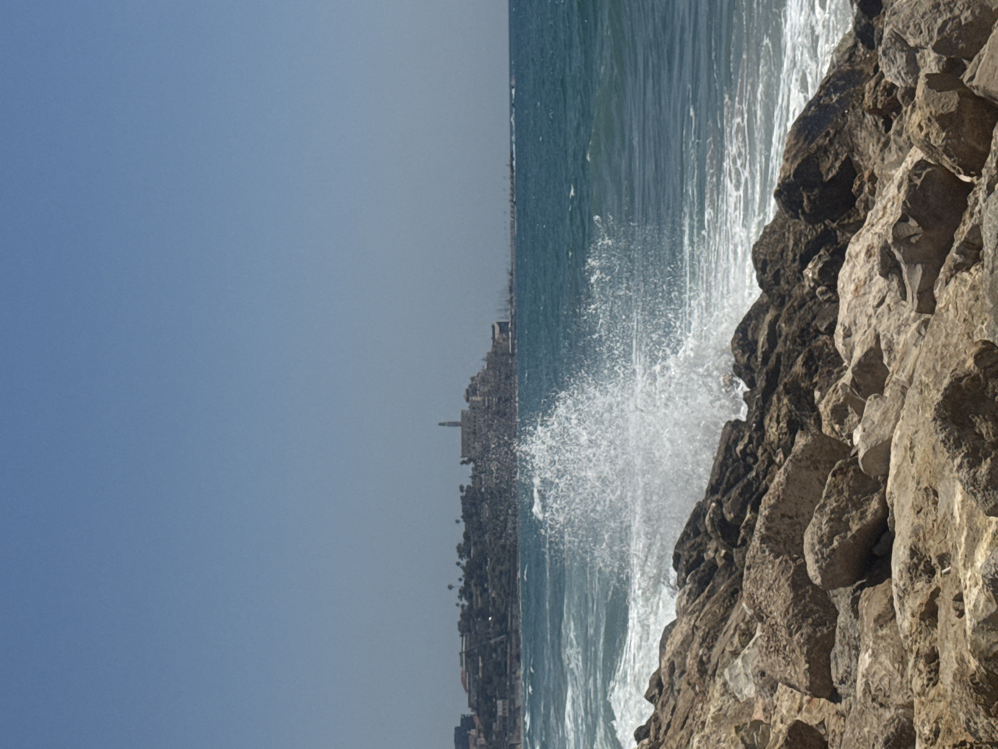 The image depicts a coastal scene in Tel Aviv, Israel. In the foreground, a rocky shore is visible, with large, weathered stones and boulders forming a natural barrier against the sea. The water is a deep blue, with white foam cresting on the waves as they crash against the rocks. A prominent splash of water erupts mid-air, captured in the moment of impact. In the background, a distant cityscape is discernible, featuring a cluster of buildings perched on a hill overlooking the sea. The architecture suggests a historical or religious significance, with at least one prominent tower or spire rising above the other structures. The sky above is clear and a light blue, indicating fair weather. The overall impression is one of a dynamic coastal environment, with the power of the sea contrasted against the enduring presence of the city.