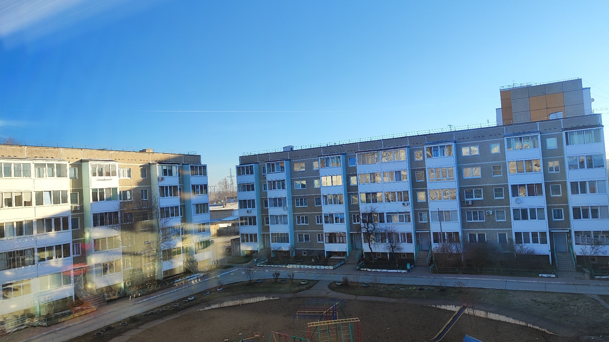 The image depicts a view from an elevated position, likely a window, overlooking a residential area in the city of Chaikovsky, Russia. Two apartment buildings dominate the scene, characterized by their Soviet-era architecture with repetitive window patterns and protruding balconies. The building on the left exhibits a lighter facade than the one on the right. In the foreground is a playground covered in dirt or sand, with several pieces of play equipment visible. The sky is clear and blue, with a faint contrail visible across the upper portion of the image. The image is taken during daylight.