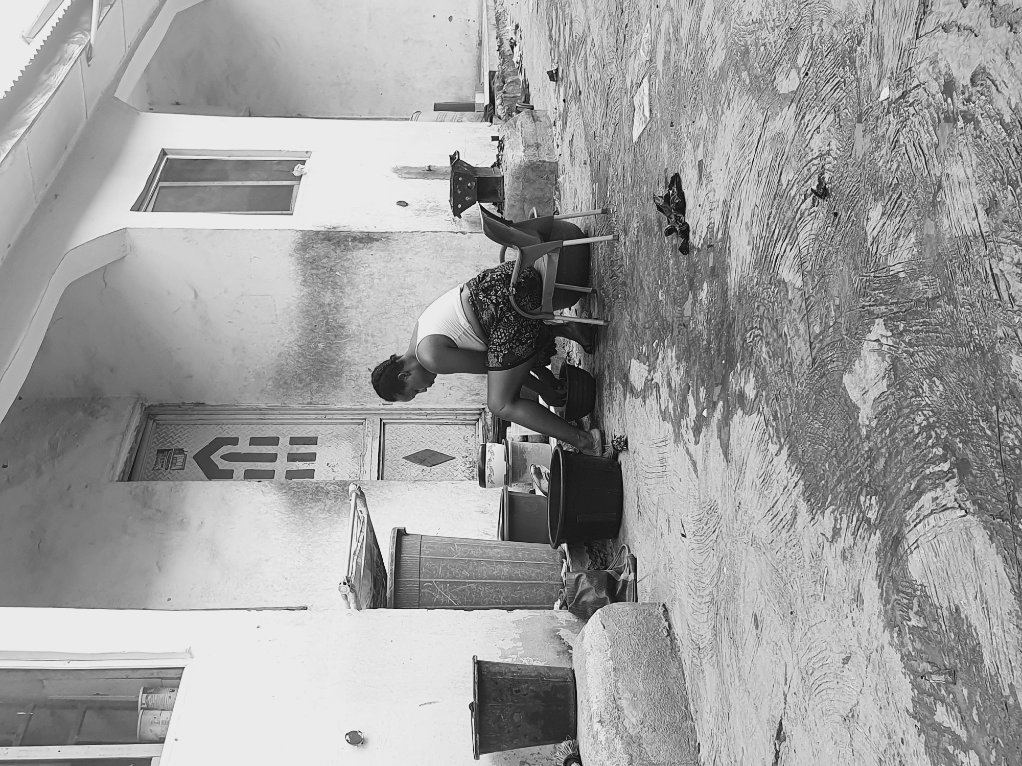 The black and white image captures a scene in Lokoja, Nigeria. A woman is seated on a chair, leaning forward with her hands inside a bucket. The setting appears to be an outdoor courtyard or alleyway adjacent to a building. There are several buckets and containers arranged in a line, suggesting the area is used for washing or cleaning. The building's wall features a door with geometric designs, a window, and what appears to be textured concrete. The ground is uneven, potentially concrete, and shows signs of wear. A pair of shoes is discarded next to the chair.