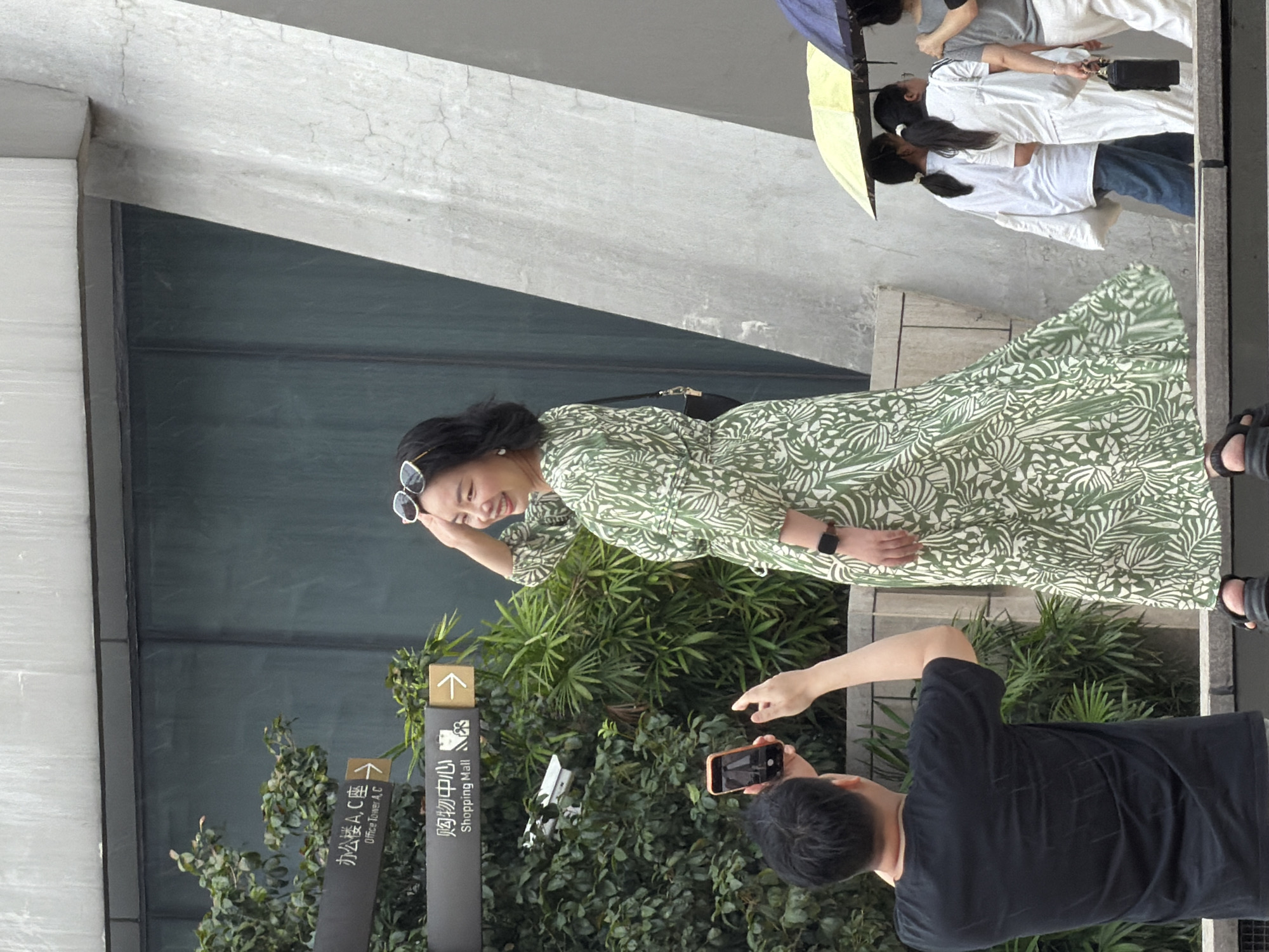 In the city of Yuzhong District, China, a woman in a green and white patterned dress poses for a photo on a tiered concrete structure. She is wearing white sunglasses pushed up onto her head, a black purse on her right hip, and black sandals. A man in a black t-shirt stands in front of her, holding a smartphone and taking her picture. Behind the woman and to the right, a group of people are walking with umbrellas, one yellow and one blue. Signage on a dark pole indicates directions to "Office Tower A, C" and "Shopping Mall." Green foliage is visible to the left of the signage and behind the man. A large, textured concrete pillar and a dark reflective windowed building form the background.
