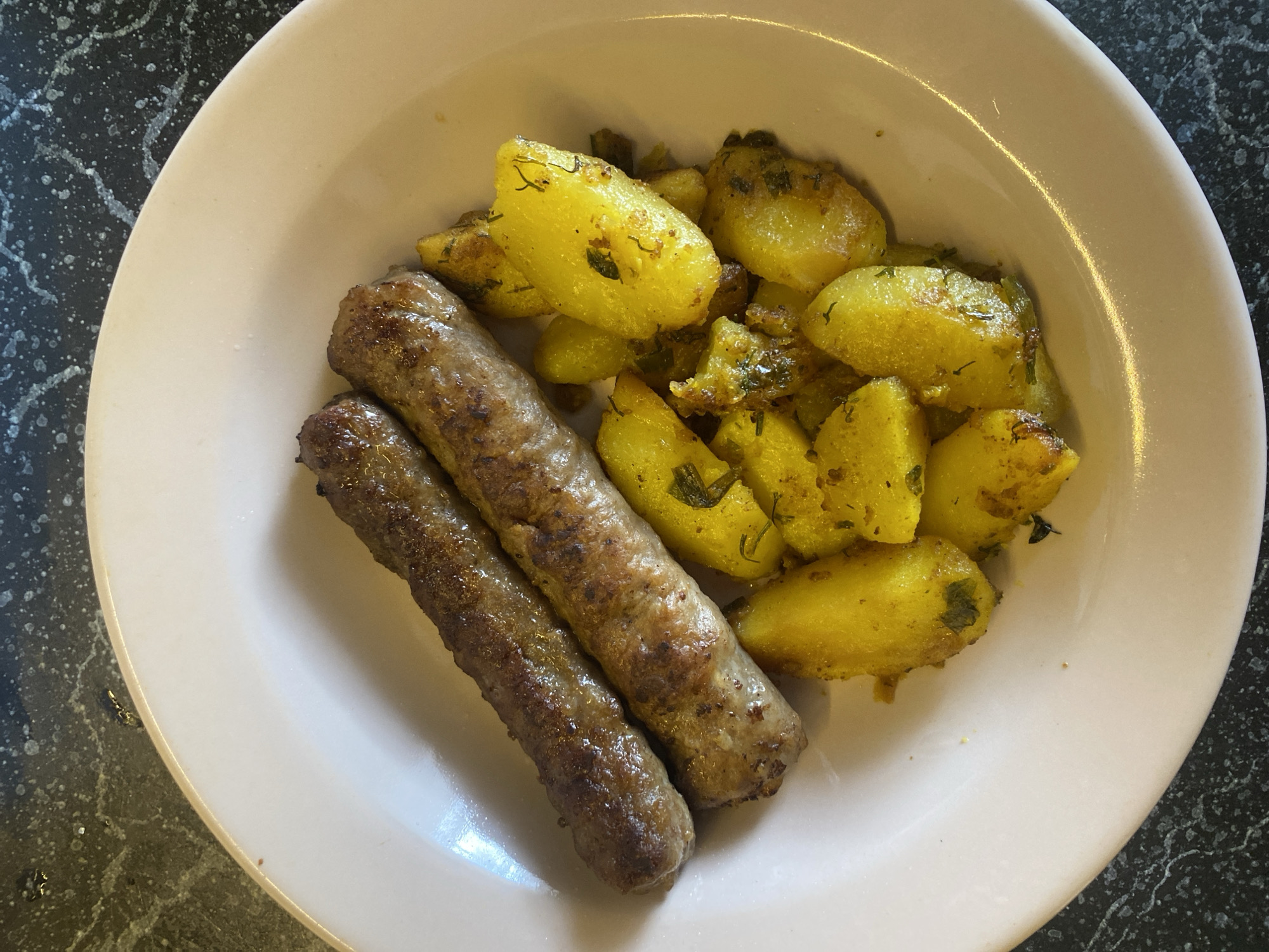 A close-up, overhead shot showcases a white plate filled with food. Two long, browned sausages lie side-by-side at the top of the plate. Beneath them, a generous portion of golden-yellow, cubed potatoes, seasoned with herbs and spices, fills the rest of the plate. The food is presented on a dark, speckled countertop. The overall scene is well-lit, highlighting the textures and colors of the meal. No city or location in Russia is identifiable from the image.