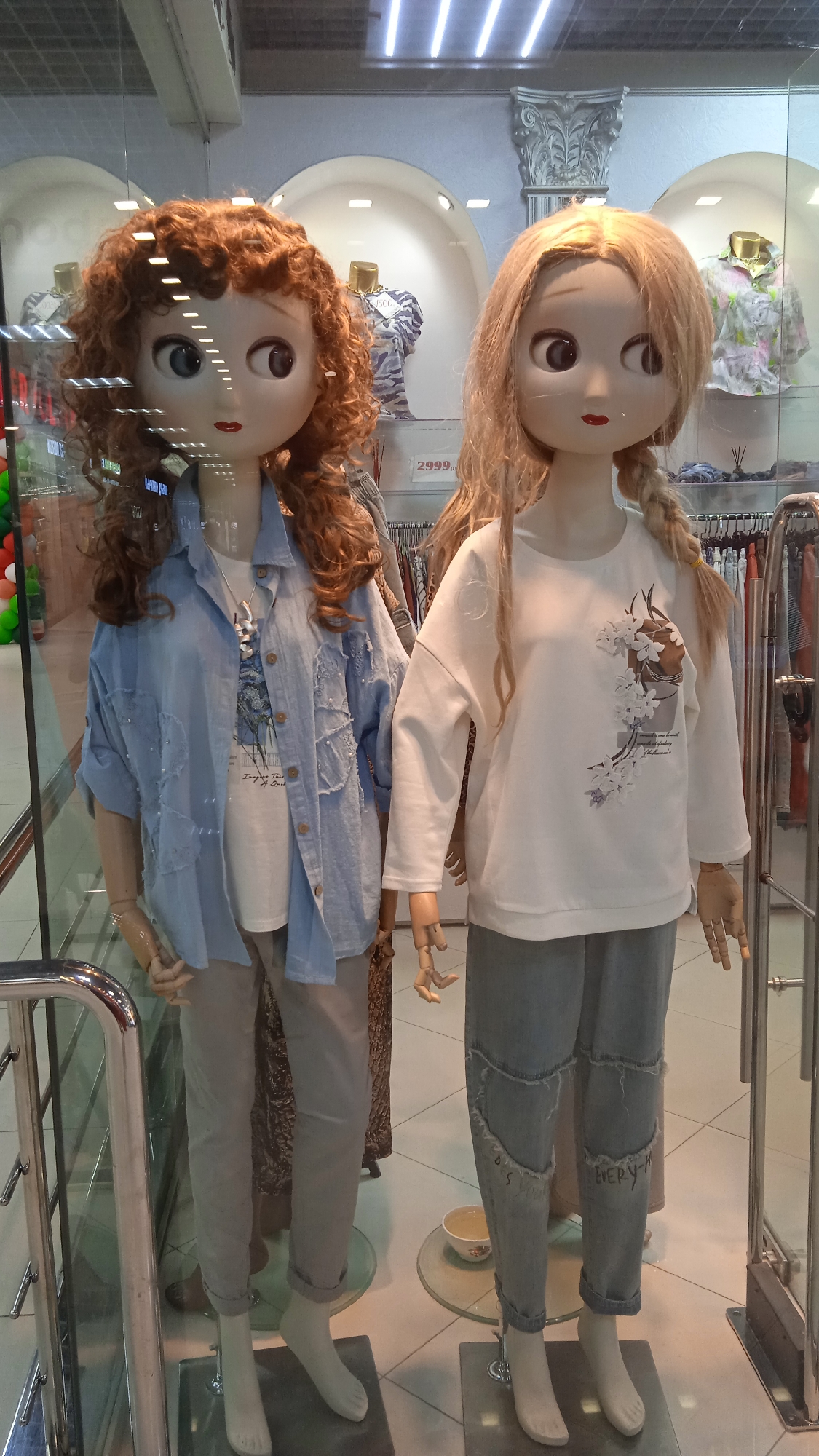 The image depicts two female mannequins in a shop window display. The mannequin on the left has curly auburn hair and is wearing a light blue open denim shirt over a white t-shirt with a graphic print, and light grey pants. The mannequin on the right has straight blonde hair styled in two braids and is wearing a white long-sleeved shirt with a floral graphic print and light blue distressed jeans. Both mannequins have large eyes and distinct facial features. Behind the mannequins, clothing racks and displays are visible, suggesting the mannequins are in a retail store. A price tag displaying "2999," is partially visible in the background. The shop window is made of glass, reflecting the interior of the store and the lighting from the ceiling. The flooring appears to be tiled. The scene is consistent with a fashion boutique located in a commercial area, possibly in Moscow, Russia, given the price format and the general aesthetic of retail displays.